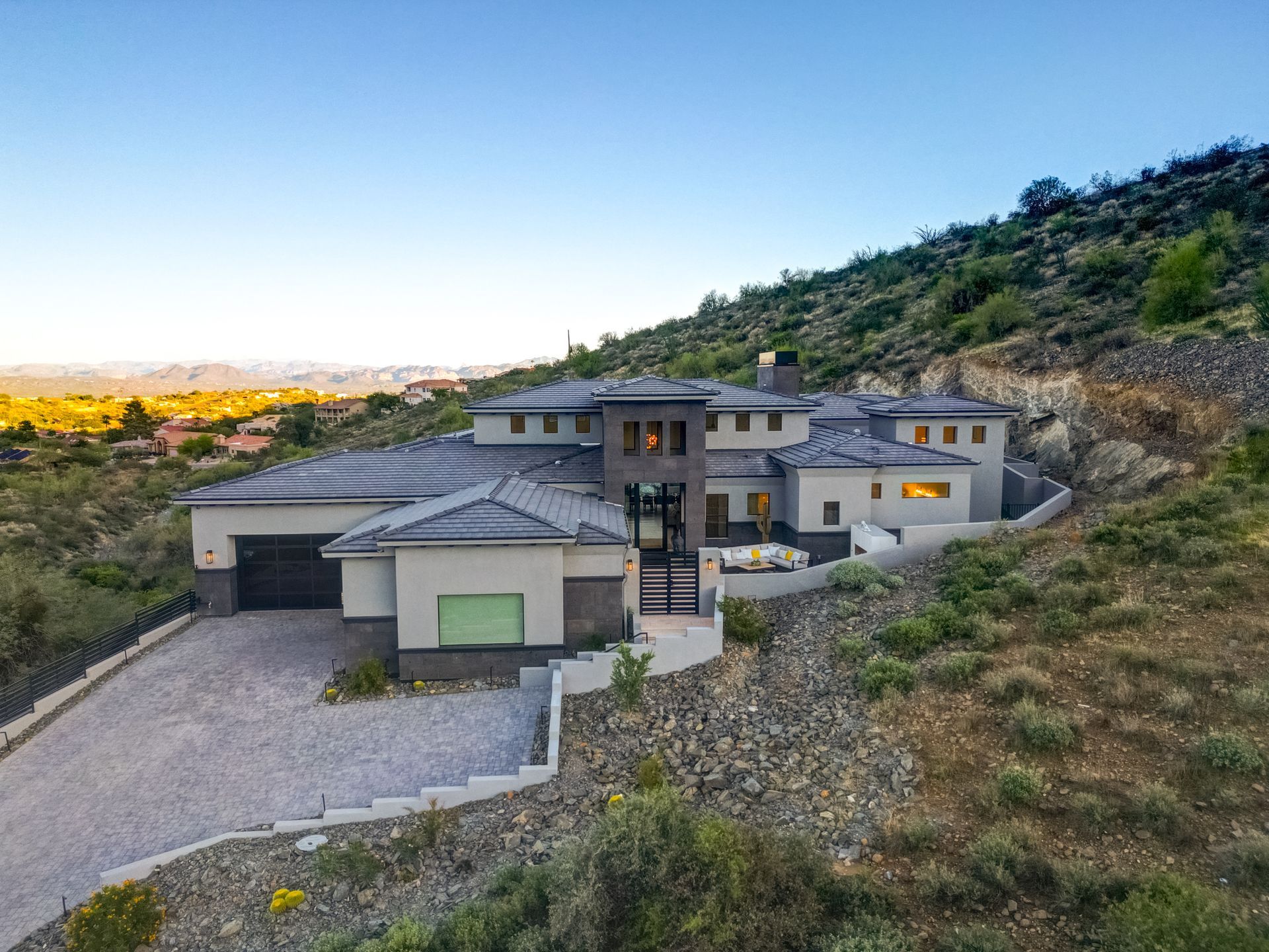 Modern home on a hillside with a gravel driveway, overlooking a distant city at dusk.
