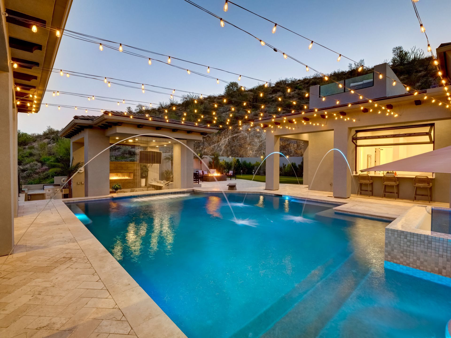 Pool with fountains, outdoor fireplace, and string lights at dusk.