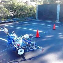 Line striping machine on asphalt, painting white lines. Cones mark off work area near black fencing.