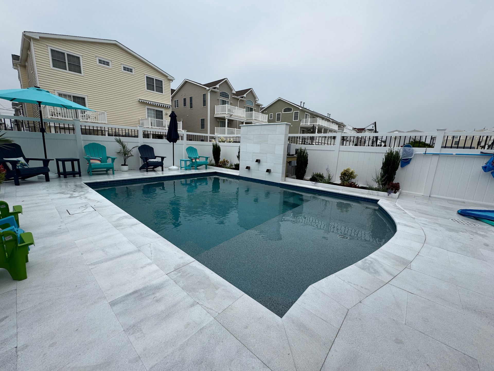Outdoor pool deck with turquoise water, white tile, lounge chairs, and buildings under a cloudy sky