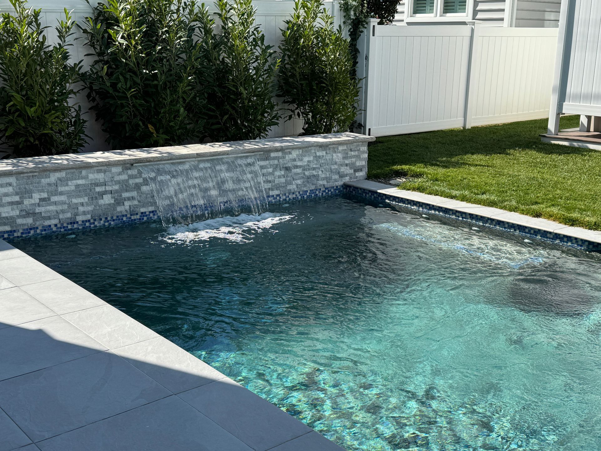 Backyard pool with turquoise water, stone wall waterfall, and green lawn beside a white fence