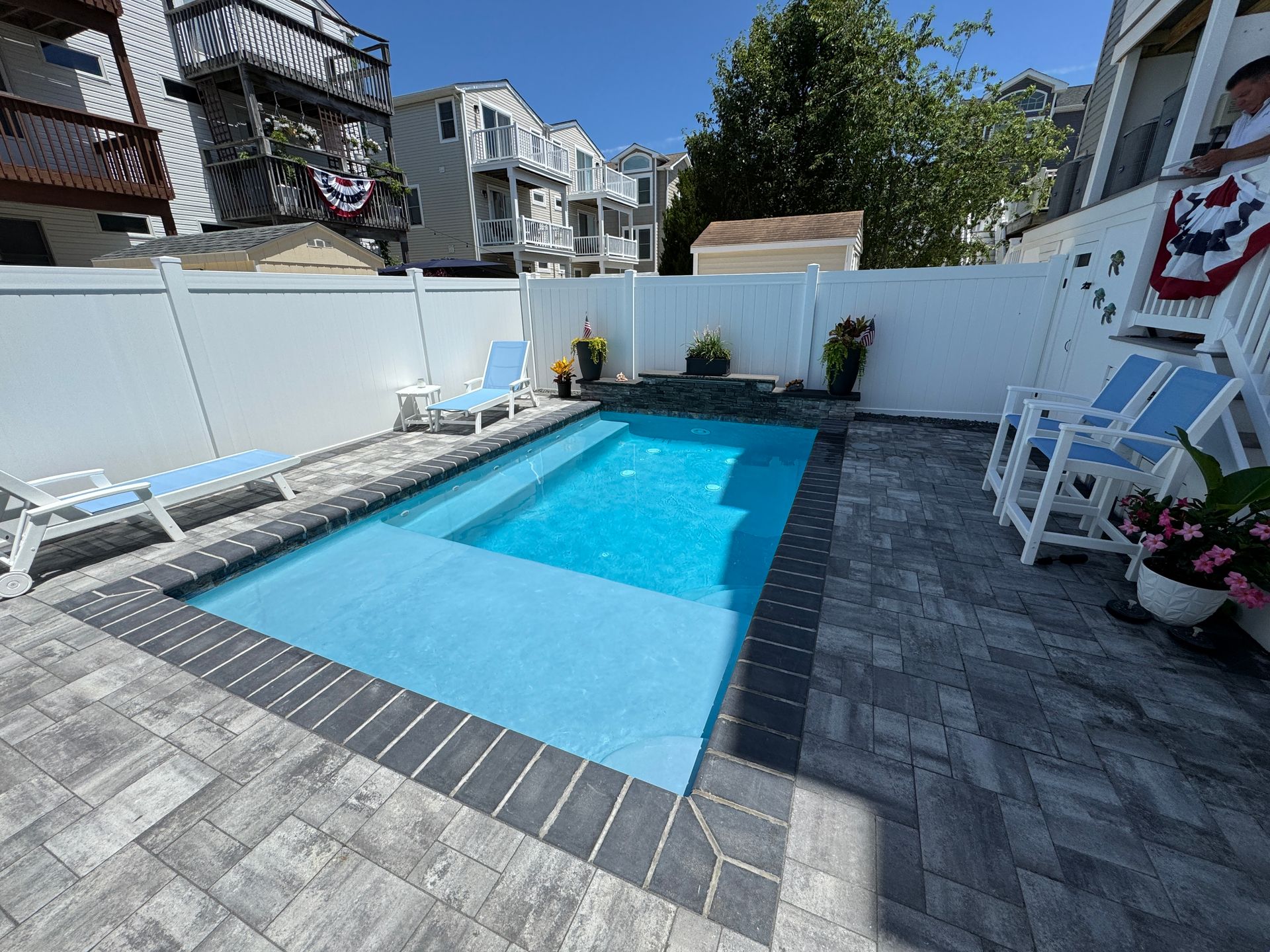 Backyard swimming pool with blue water, white lounge chairs, and patio pavers by a white fence