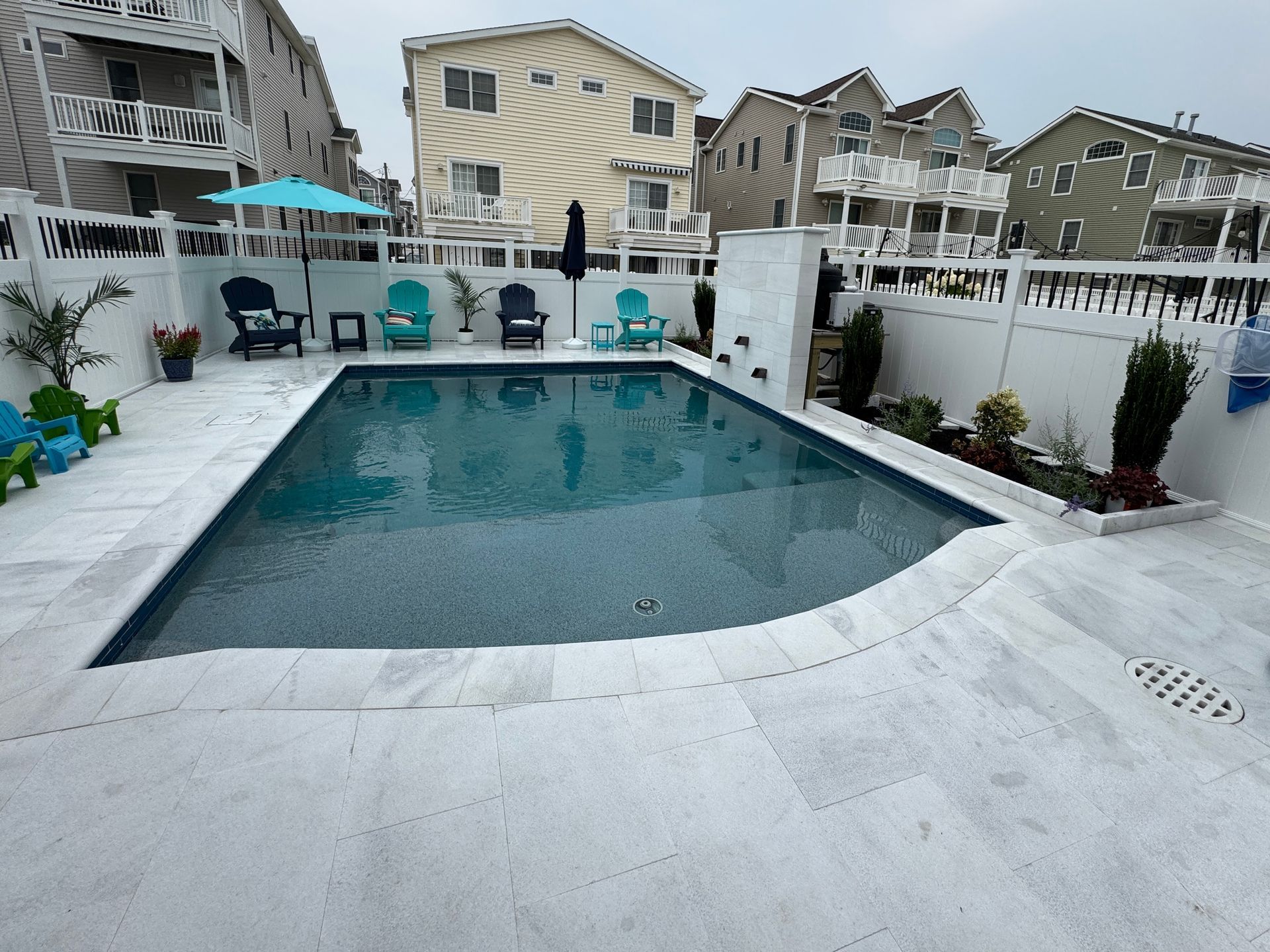 Backyard swimming pool with white deck, lounge chairs, umbrellas, and houses in the background