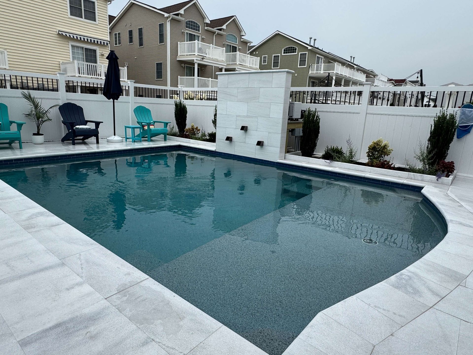 Backyard swimming pool with white deck, teal chairs, and townhouses in the background