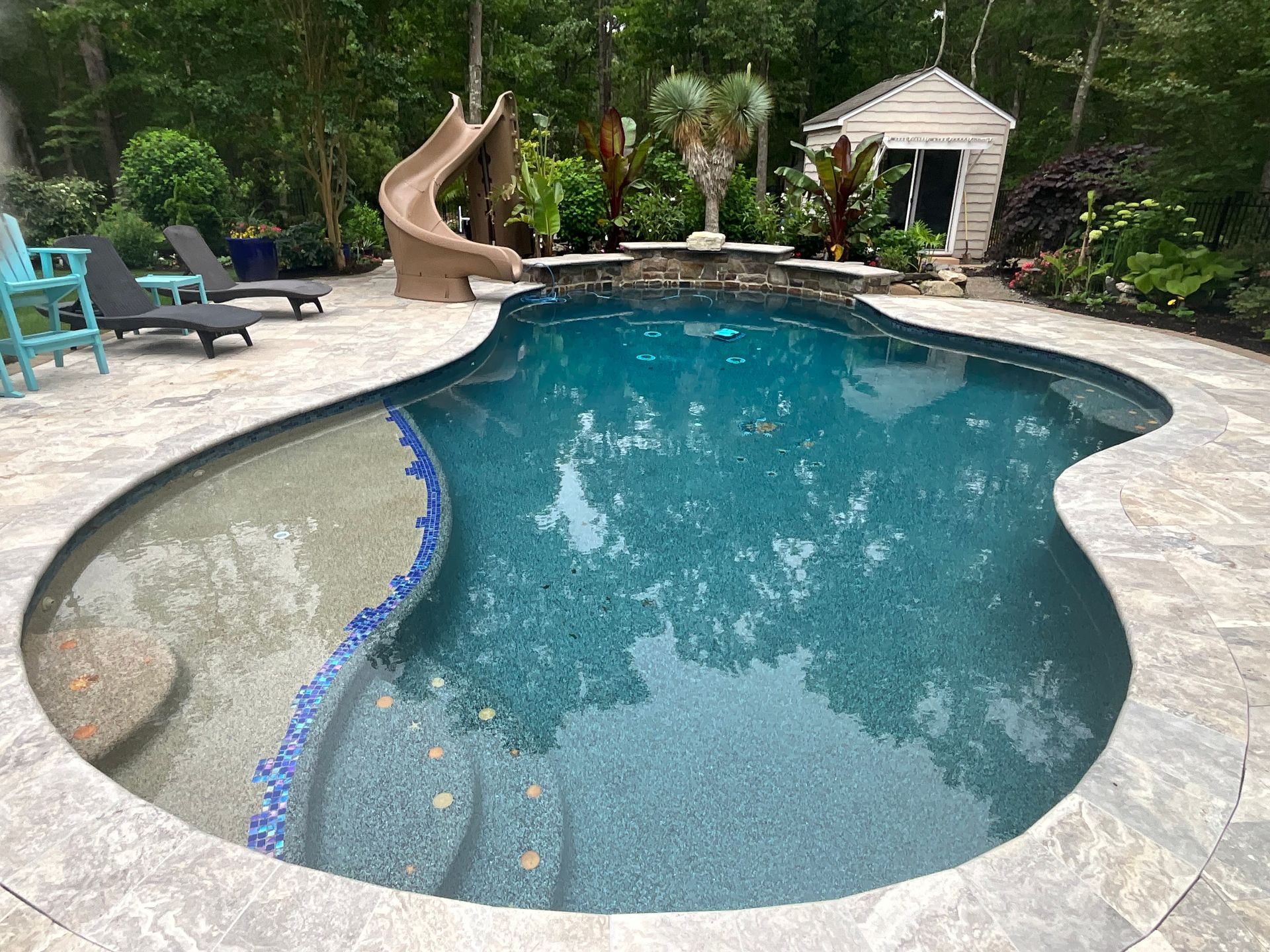 Backyard swimming pool with curved stone deck, blue water, lounge chairs, and a small gazebo and slide