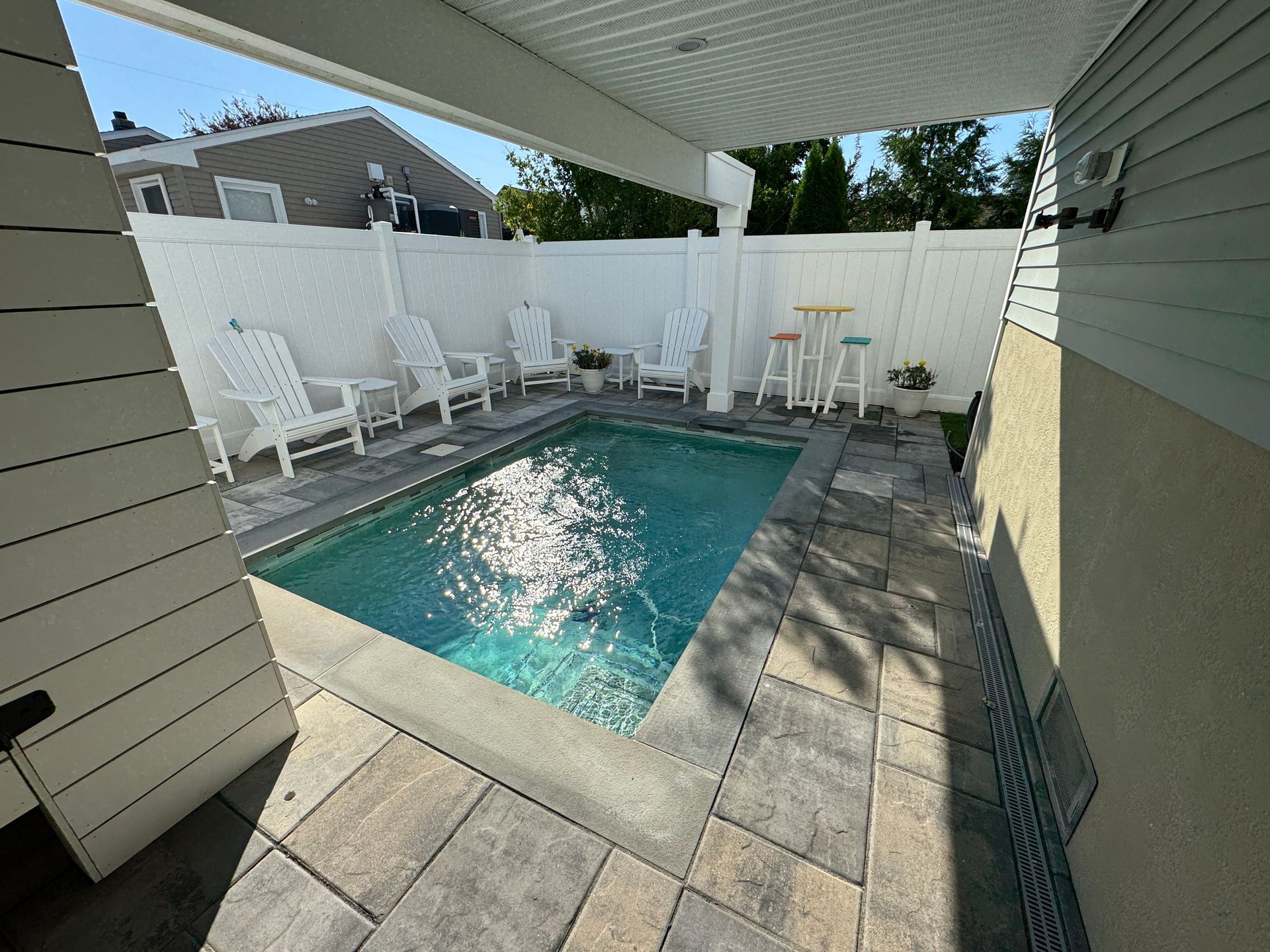 Small backyard pool with patio pavers, white lounge chairs, and a privacy fence in bright sunlight