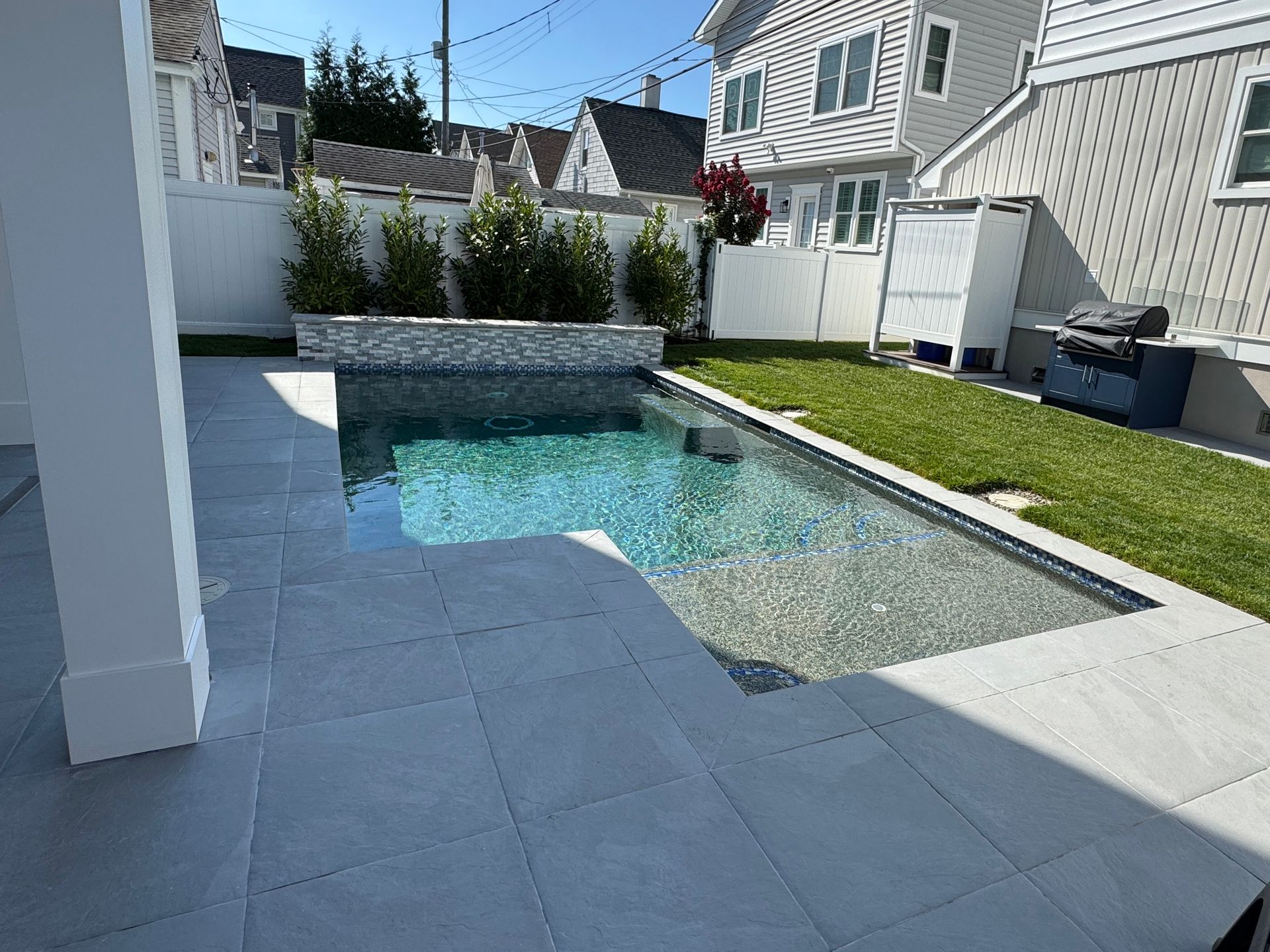 Backyard patio with a swimming pool, stone paving, green lawn, and neighboring houses under bright sun