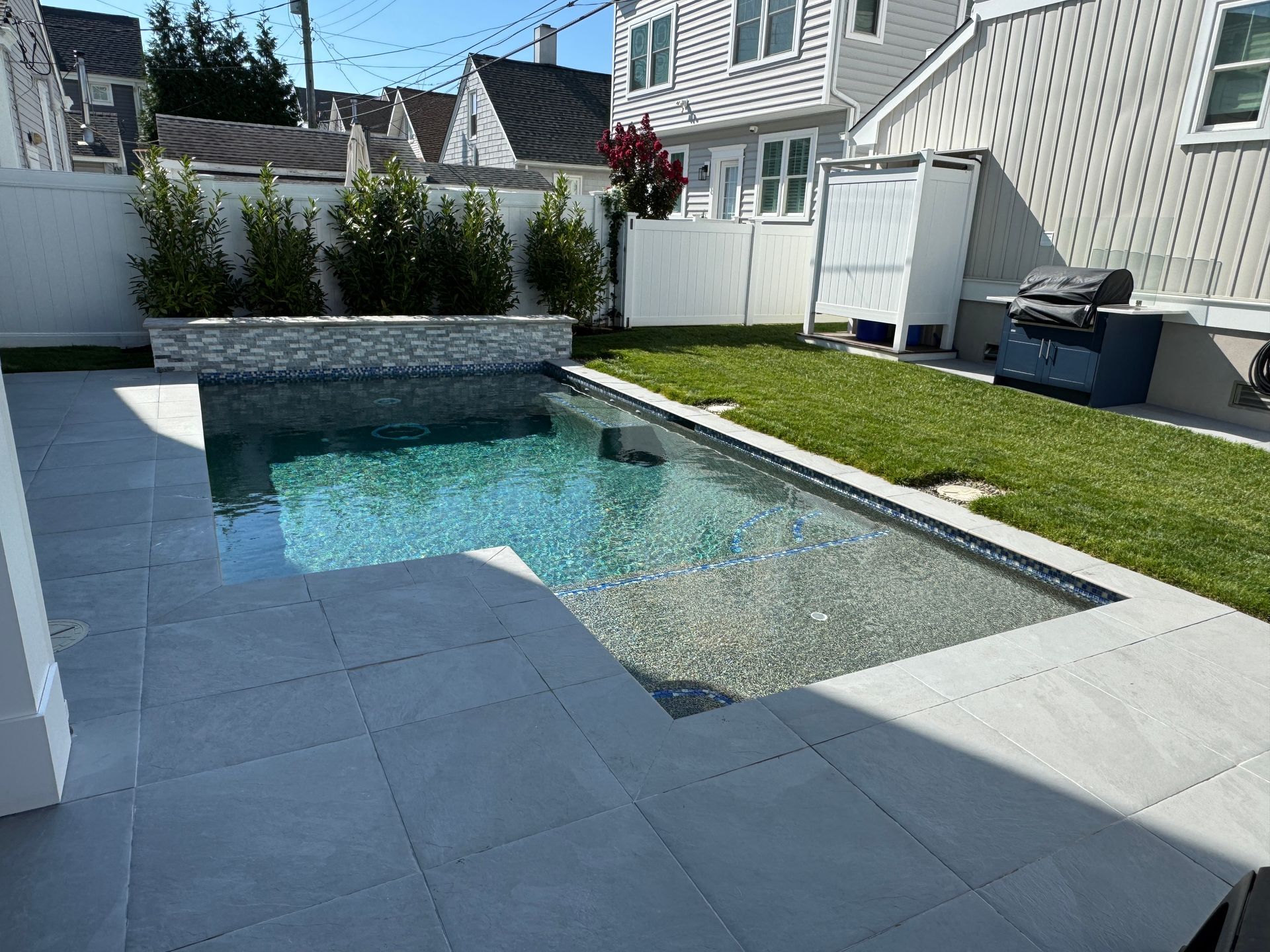 Modern backyard with a small rectangular pool, gray stone patio, and green lawn beside a house.
