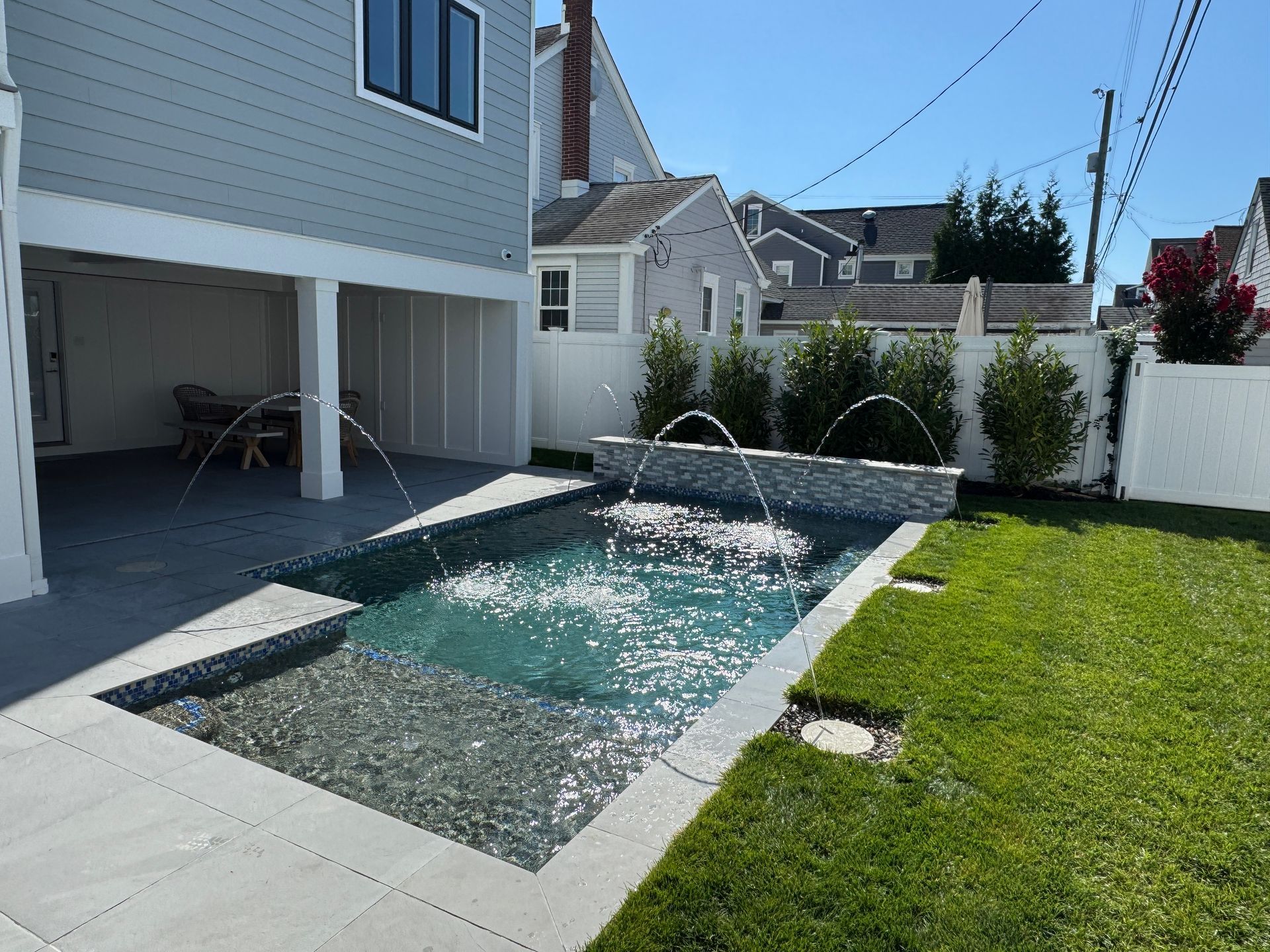 Backyard patio with a small rectangular pool, fountain jets, lawn, and a white house garage