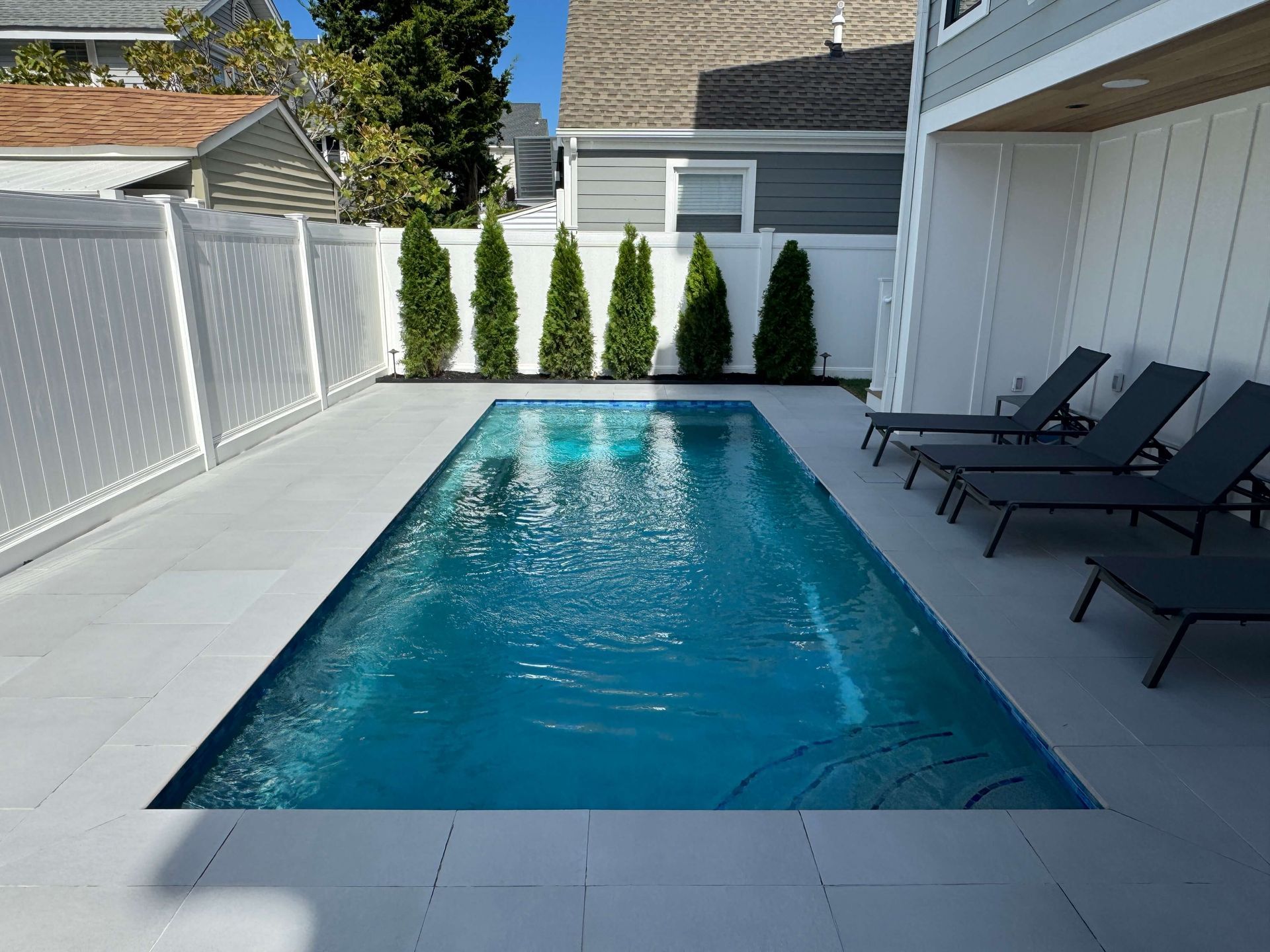 Long, narrow backyard pool beside a white fence and black lounge chairs in a sunny courtyard