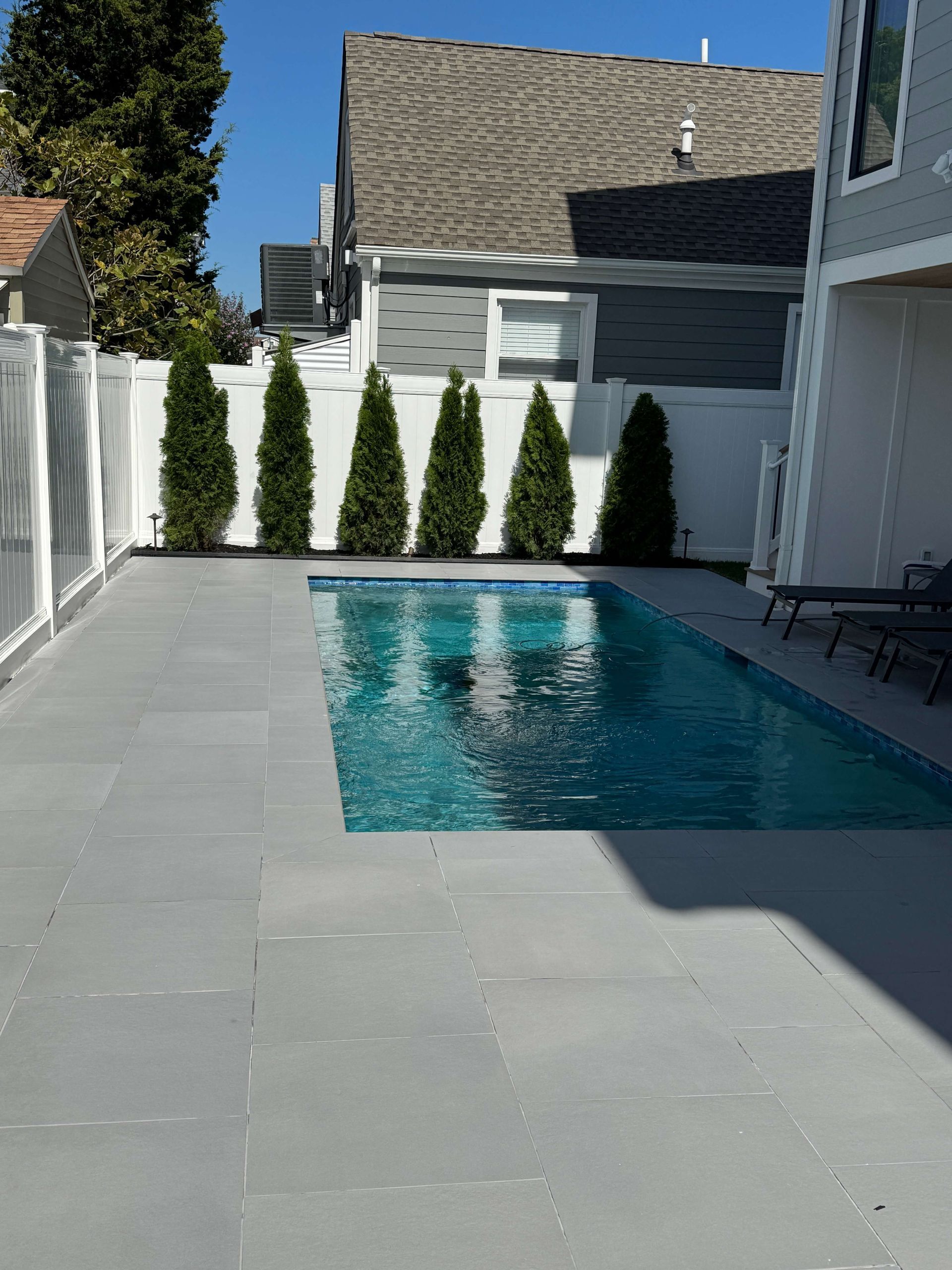 Modern backyard with a small blue pool, gray patio tiles, white fence, and tall evergreens under a clear sky