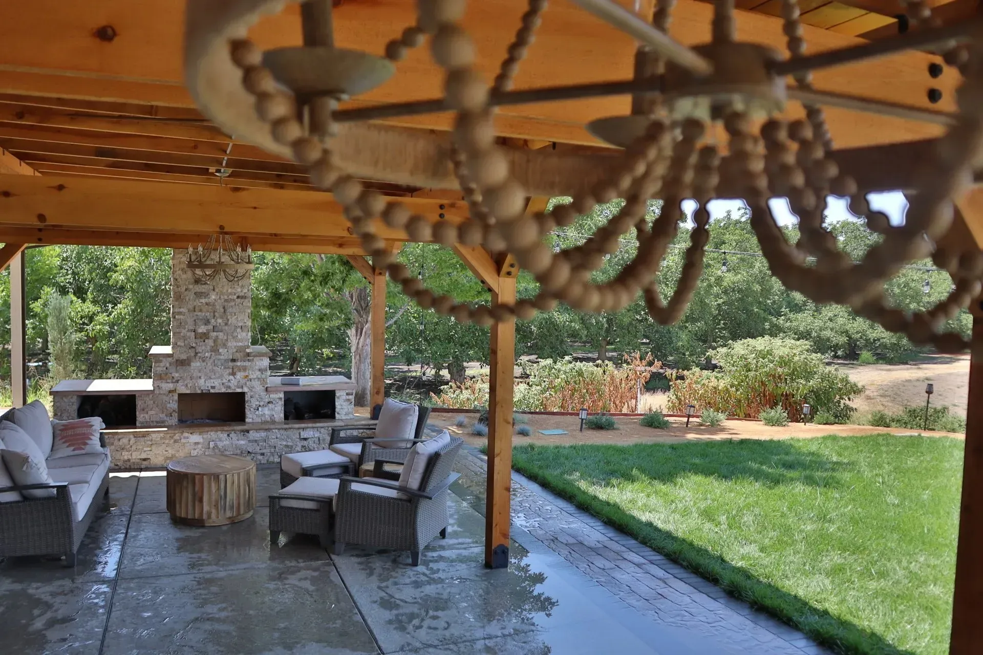 Covered patio with seating, fireplace, and chandelier overlooking a lawn and trees.