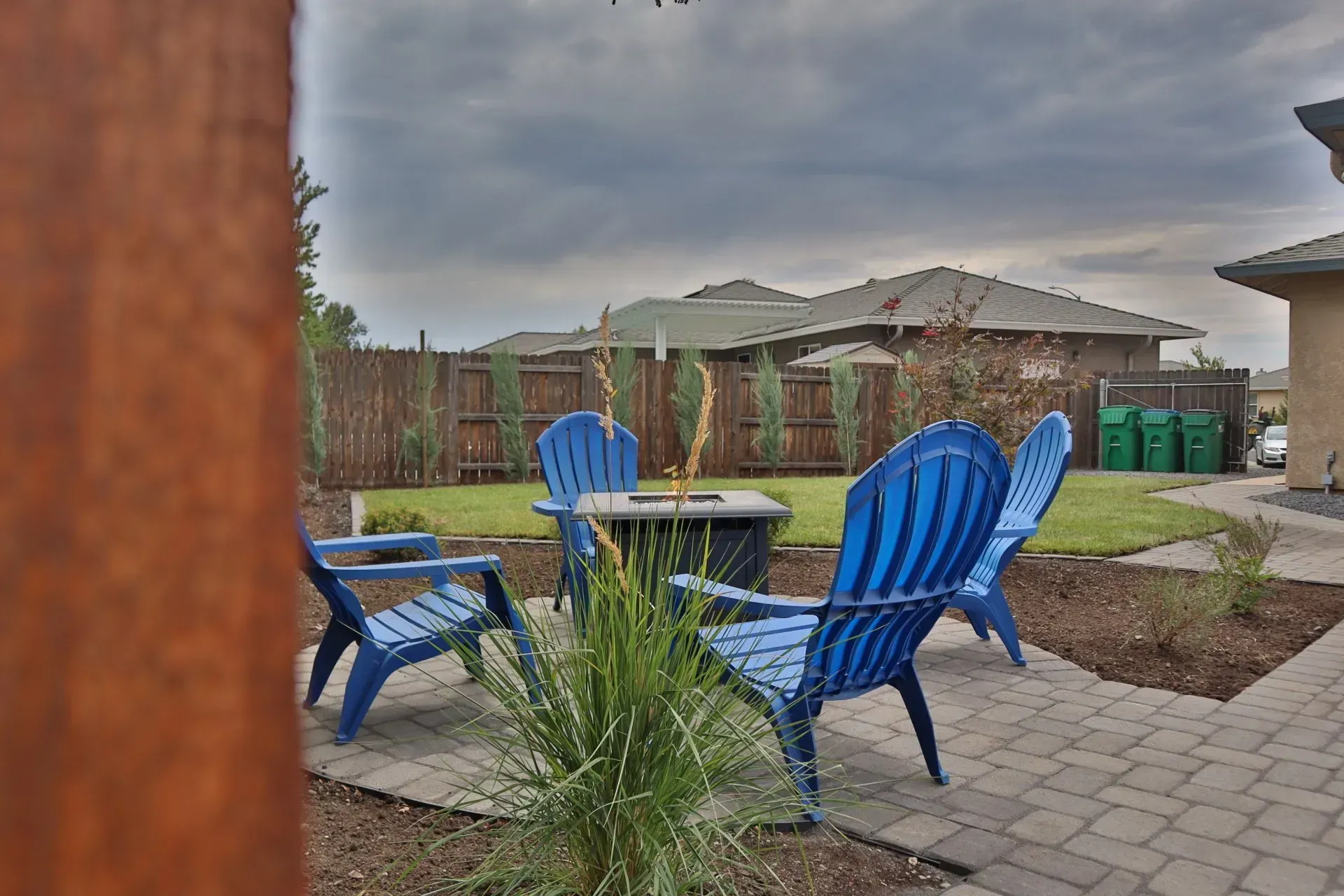 Blue chairs around a fire pit in a backyard, with a fence and houses in the background under a cloudy sky.