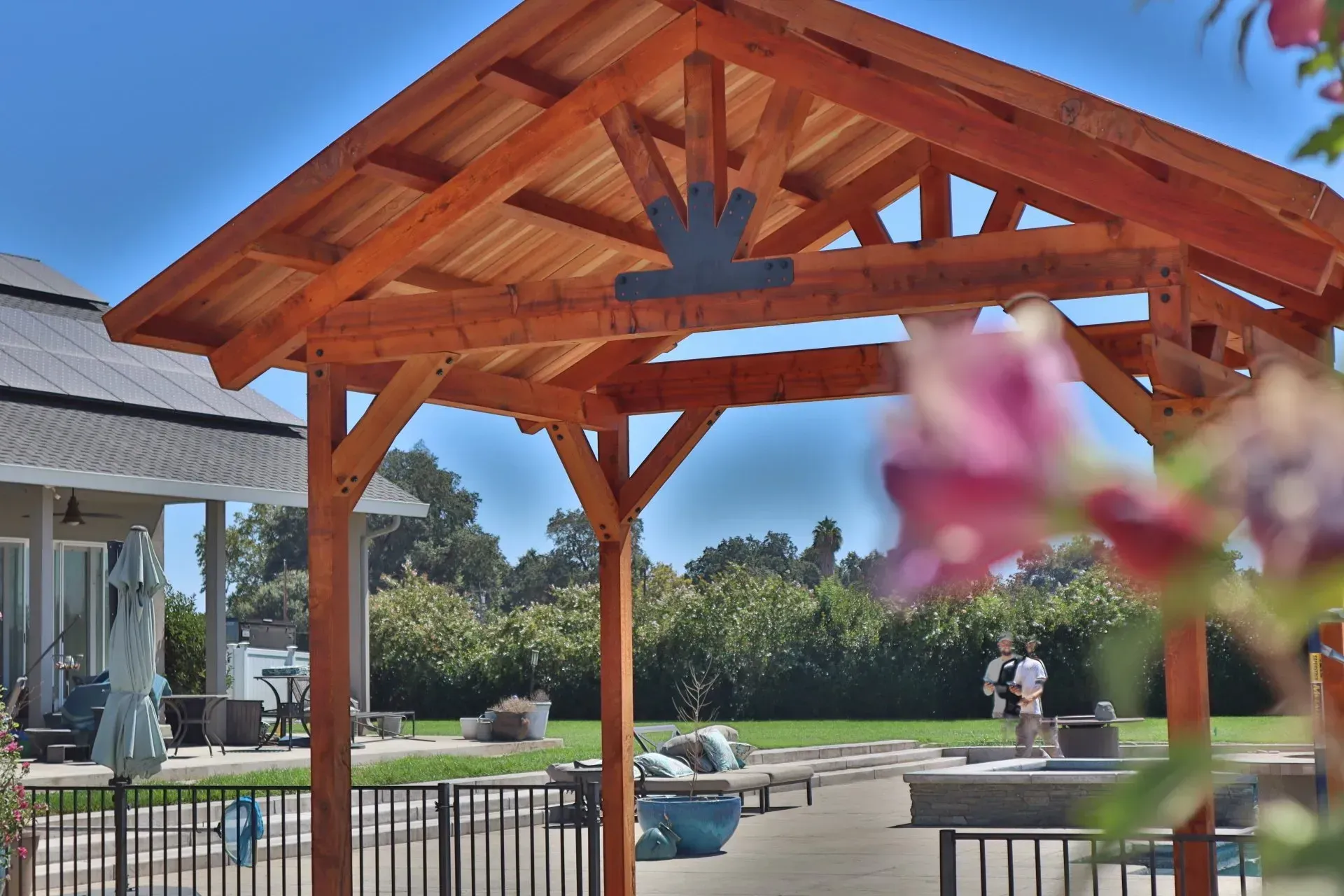 Wooden gazebo with decorative metalwork over a patio, lawn, and house on a sunny day.