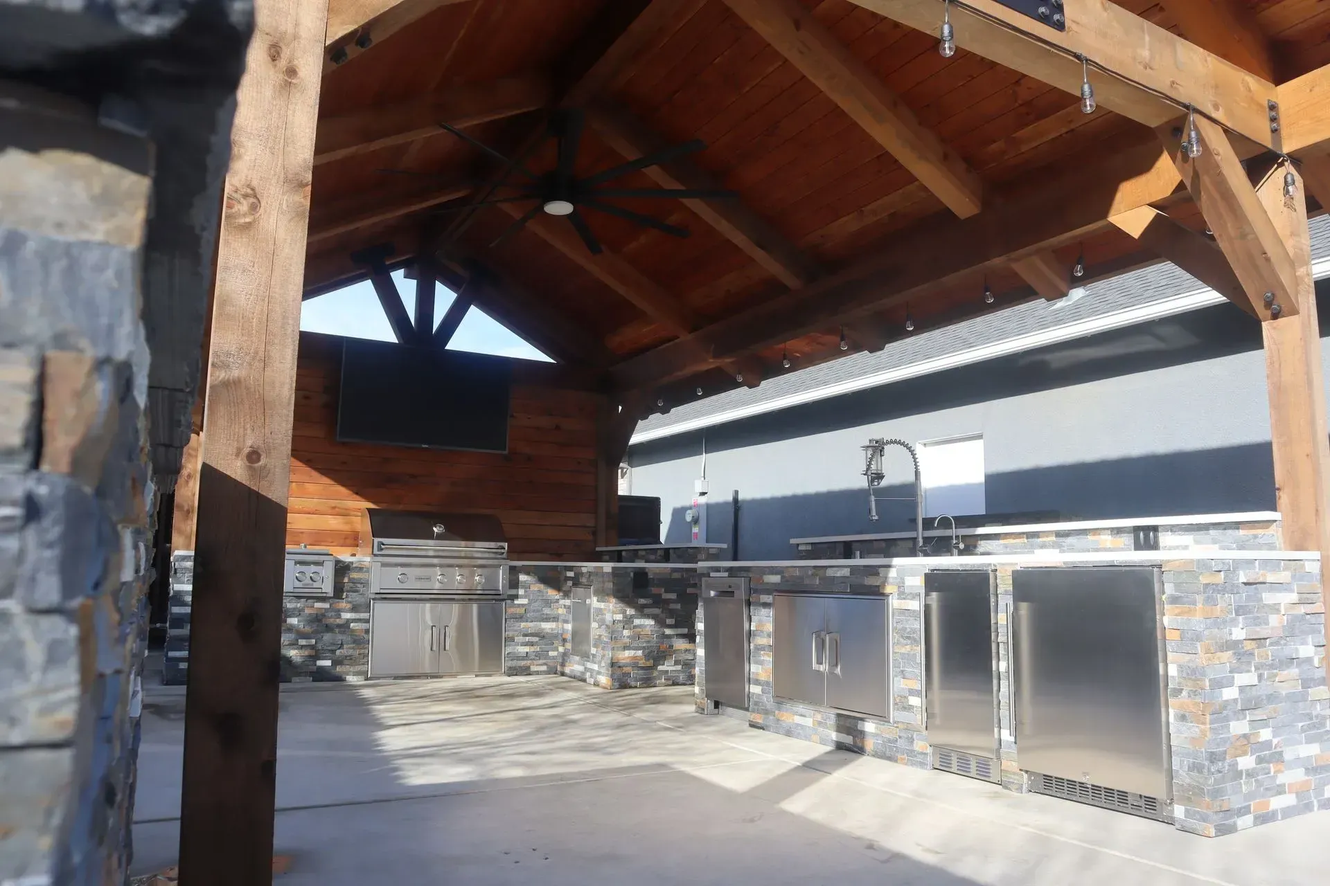 Outdoor kitchen with stainless steel appliances under a wood-beamed roof and stone accents.
