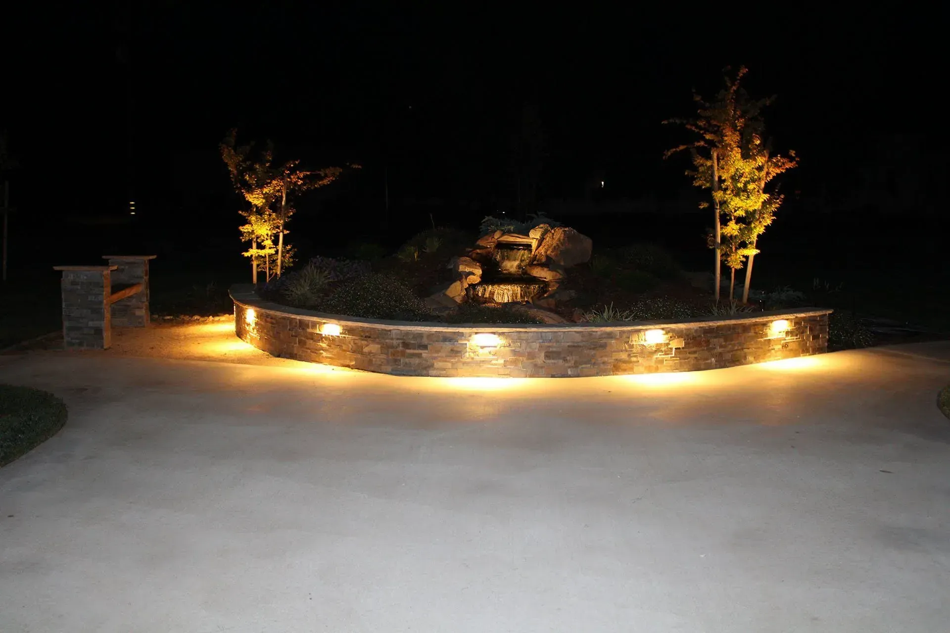 Nighttime view of a lit stone wall and a small waterfall with illuminated trees.