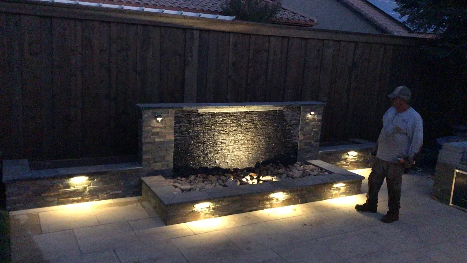 Water feature illuminated by lights in a backyard. Man standing to the side. Wooden fence in background.
