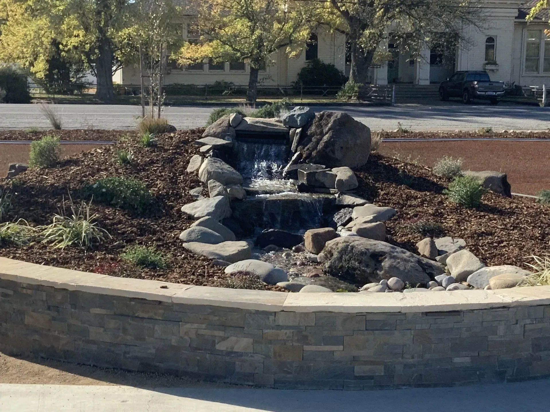 Stone waterfall feature in a circular bed with plants and a stone retaining wall in front of a street.