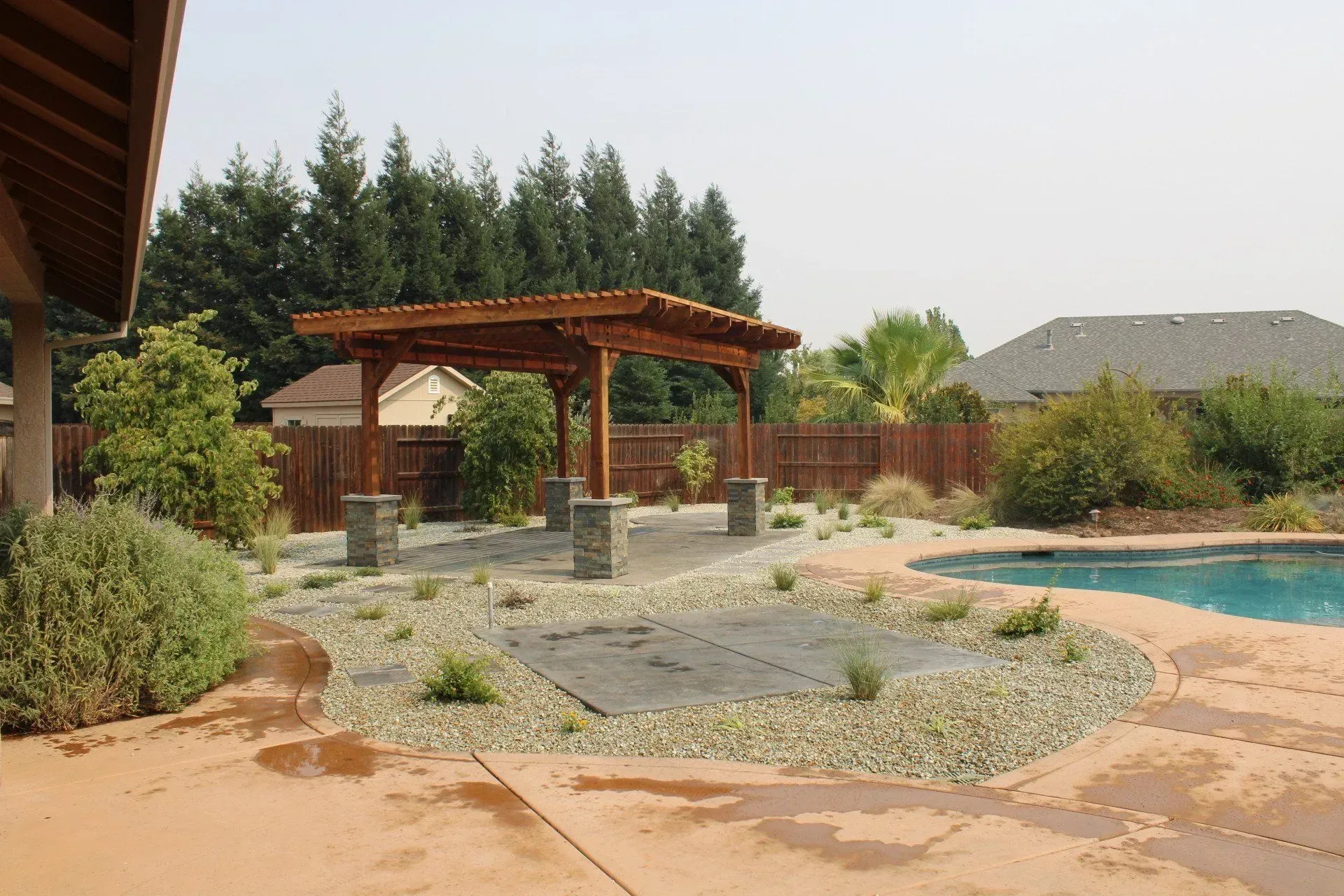 Wooden pergola over gravel area next to a pool in a backyard with trees.