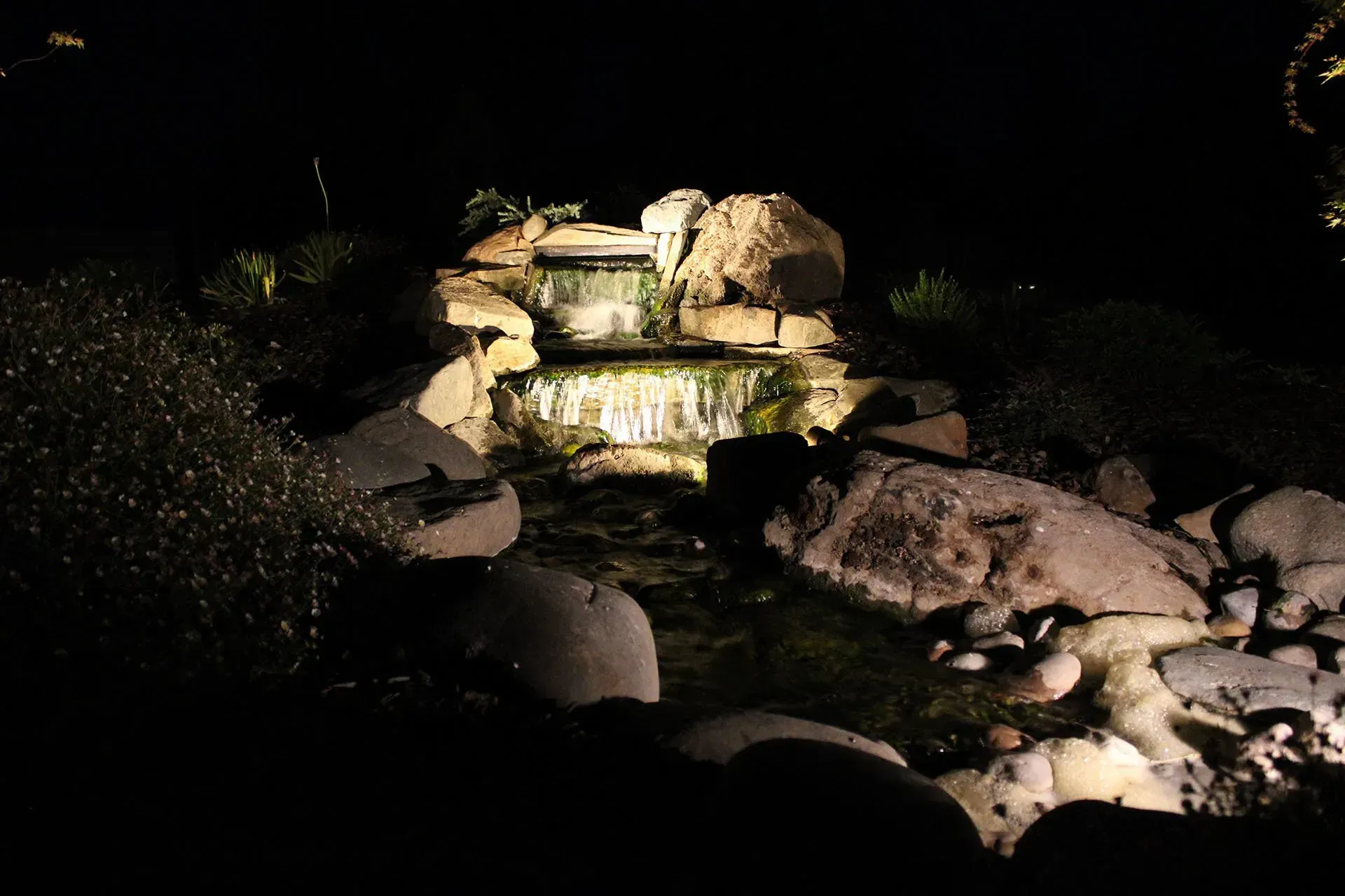 Waterfall illuminated at night, cascading over rocks, surrounded by dark foliage.