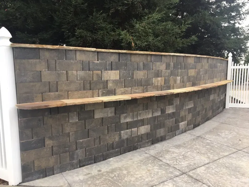 Stone wall with wooden bar top. White fence on either side; concrete patio.