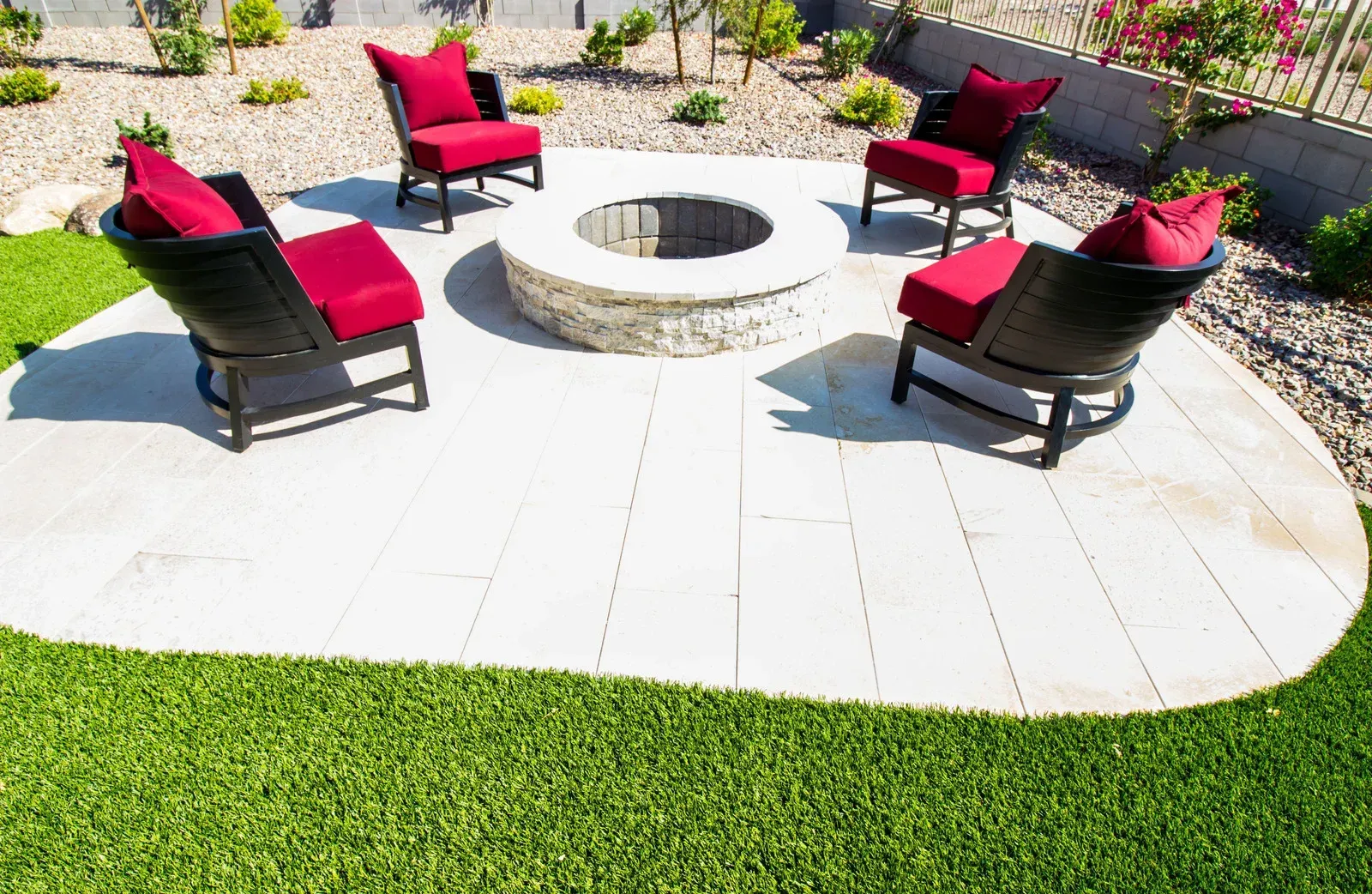 Four black chairs with red cushions around a stone fire pit on a white patio. Green grass surrounds.