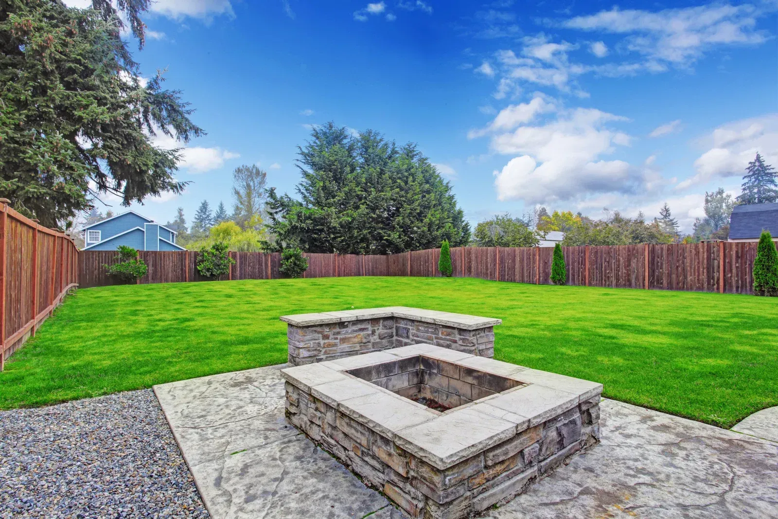 Backyard with fire pit, green lawn, brown fence, and blue sky.
