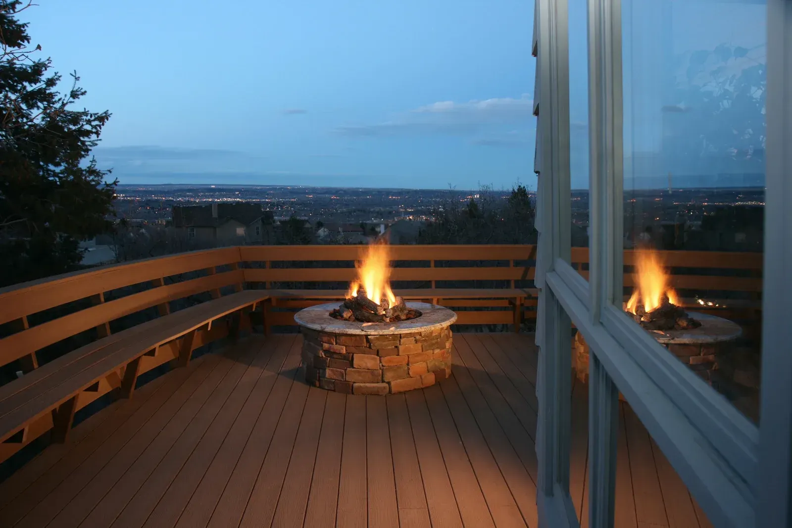 A fire pit with flames on a deck at dusk, with built-in benches and city lights in the distance.