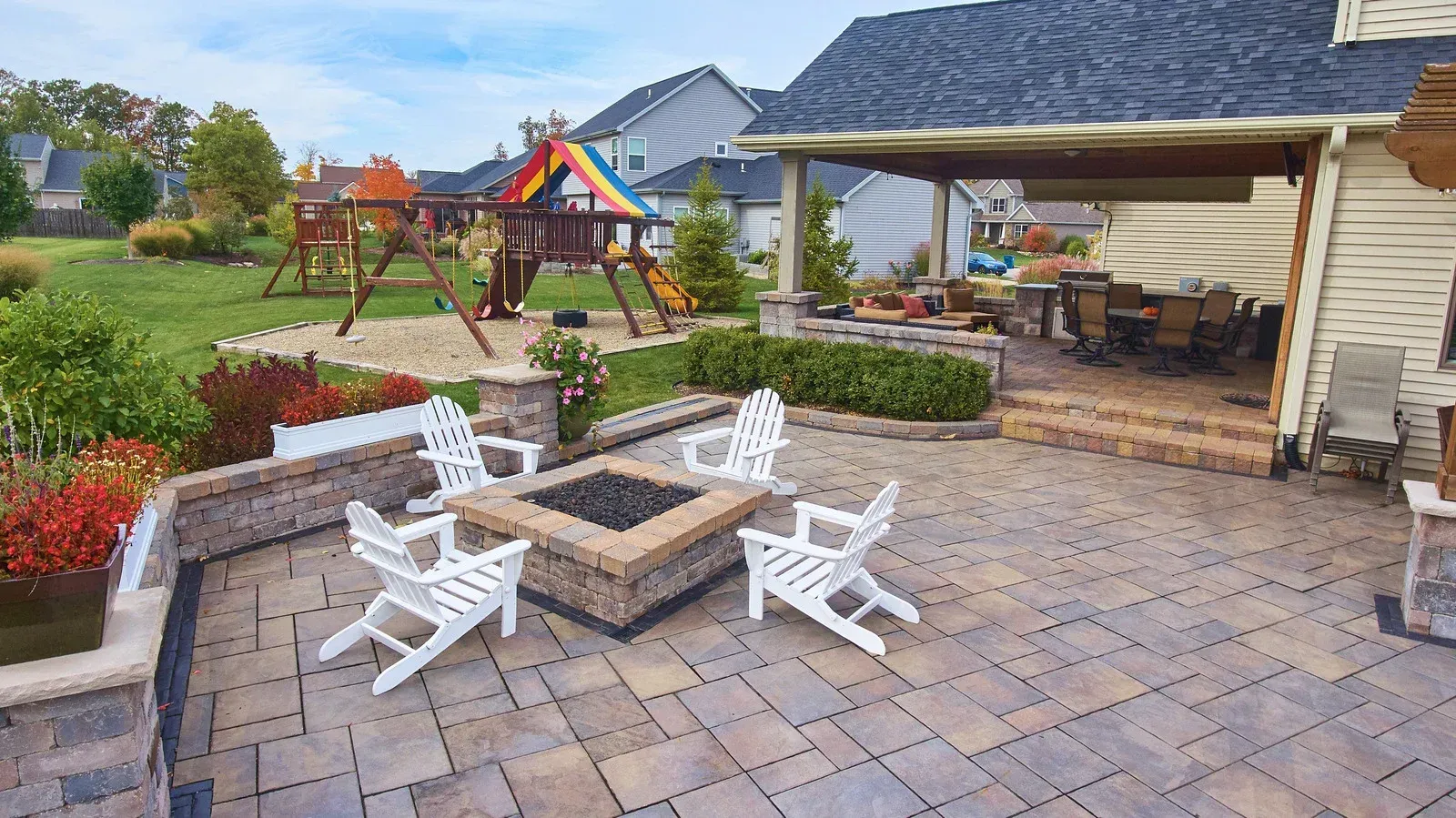 Backyard patio with fire pit, white chairs, and a play set in the background.