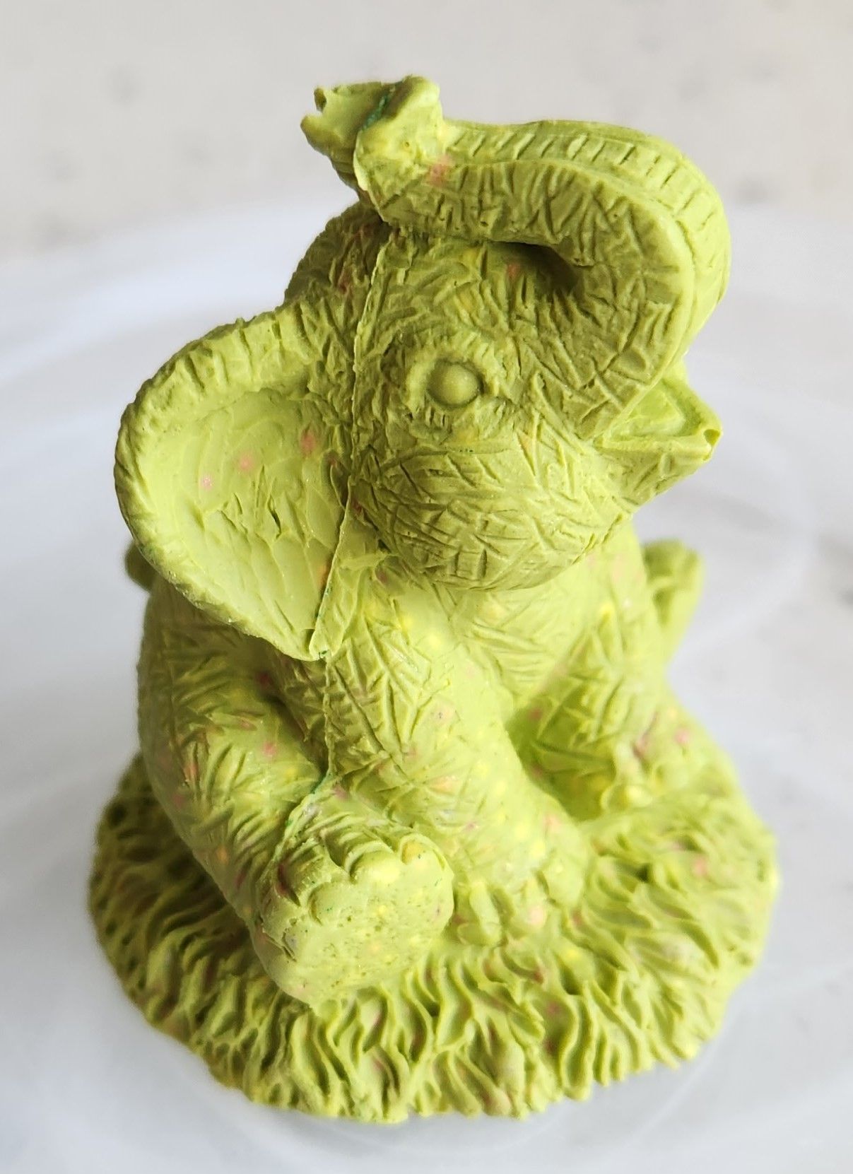 Green elephant figurine with trunk raised, sitting on grass.