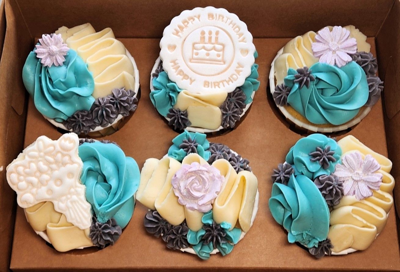 Six cupcakes with teal and cream frosting and decorative toppings in a box.