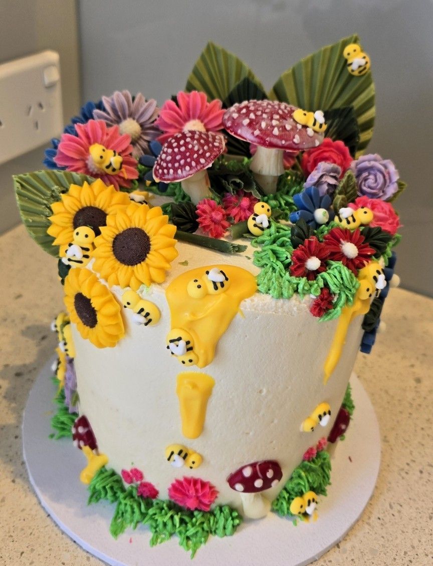 Cake decorated with colorful flowers, mushrooms, and bees; white frosting with yellow honey drips.