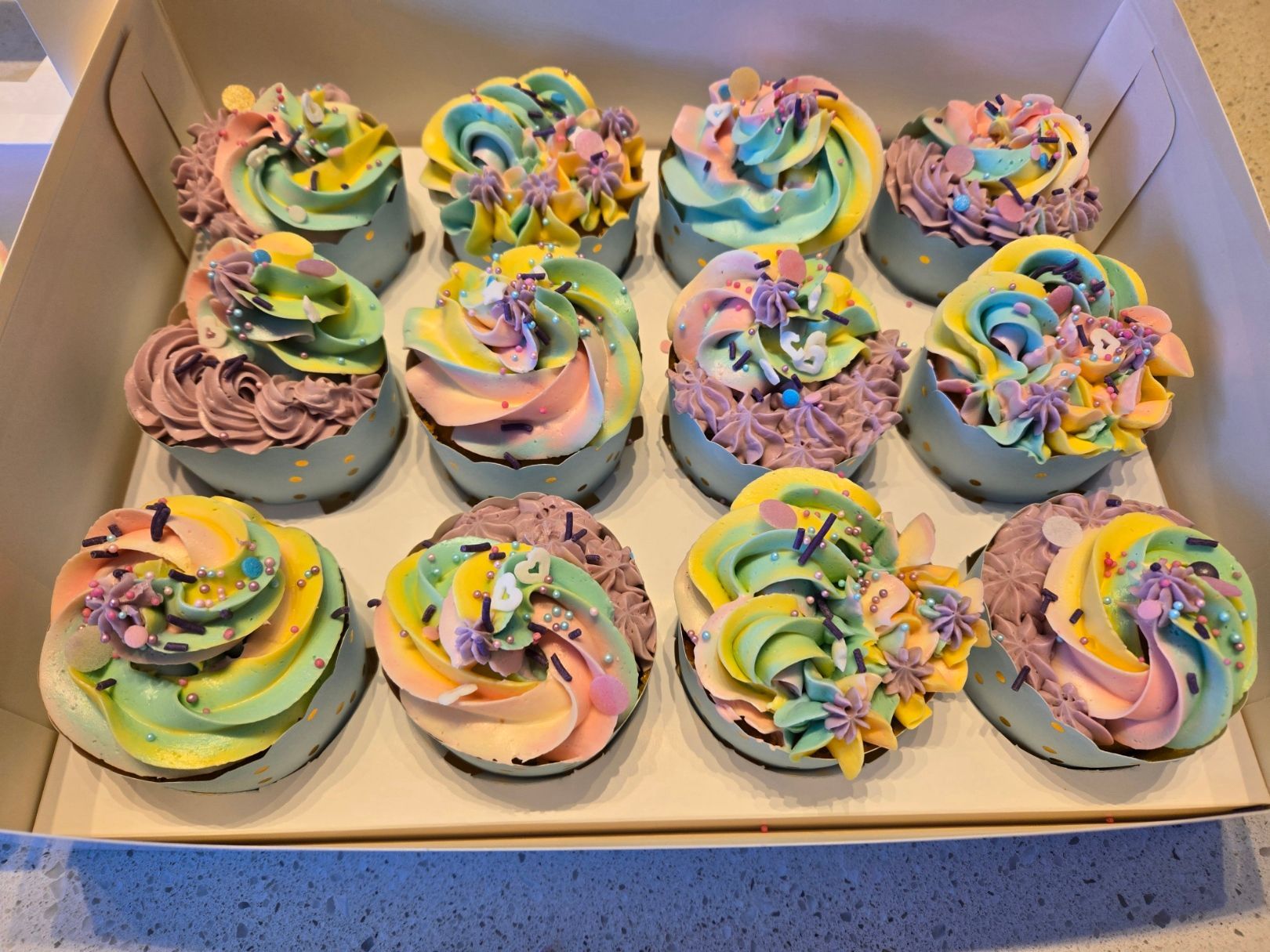 Box of colorful frosted cupcakes. Each has a different swirl of pastel frosting.