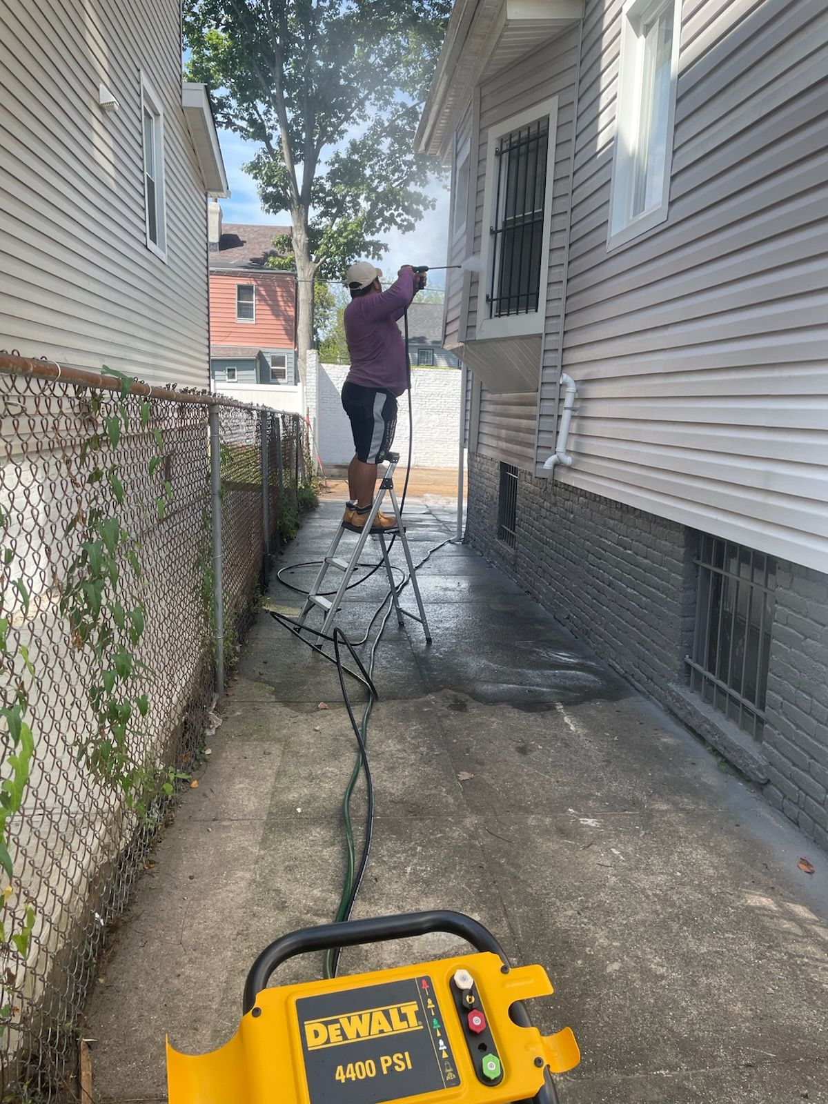 Person pressure washing a house exterior from a ladder in a narrow alleyway. A DeWalt power washer is in the foreground.