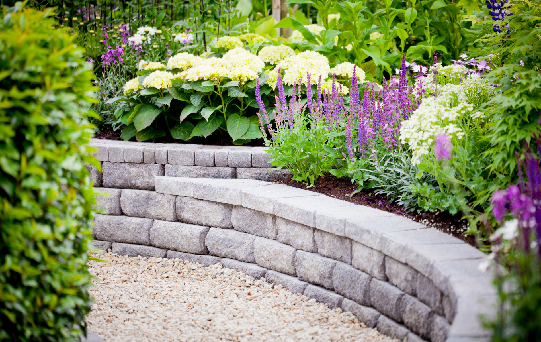 A stone wall in a garden surrounded by flowers and bushes.