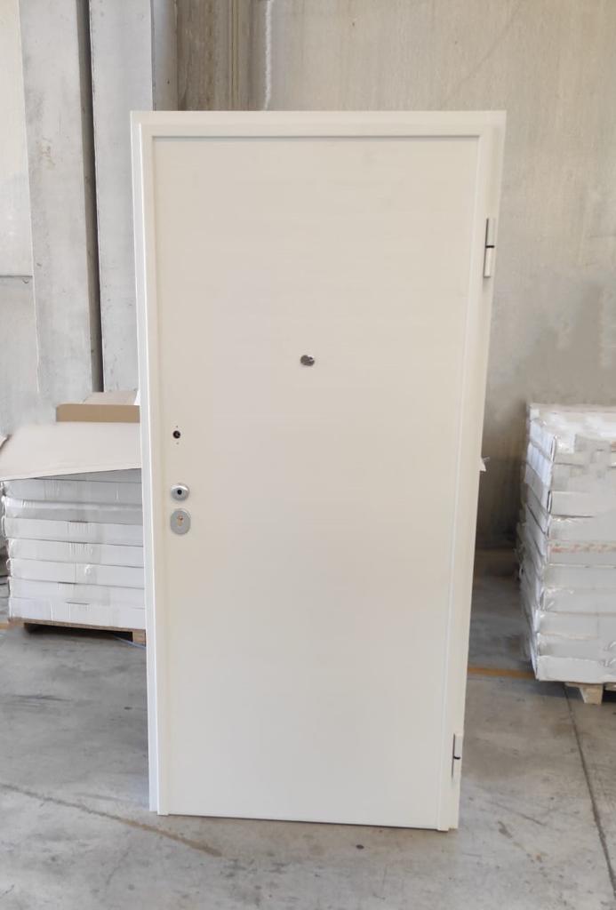 Porta in acciaio bianco con maniglia e spioncino, posta all'interno accanto a pile di materiali.