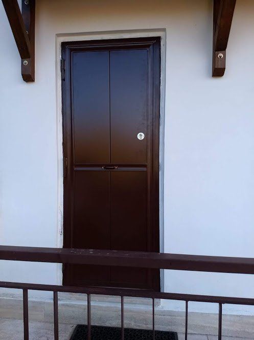 Porta marrone su una parete bianca, con una ringhiera in primo piano. Travi di legno in cima.