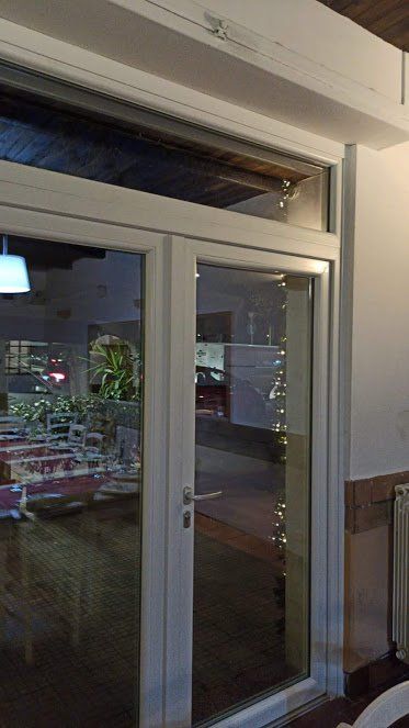 Porta a vetri di un ristorante che riflette i tavoli interni. Cornice bianca, luci lungo la finestra.