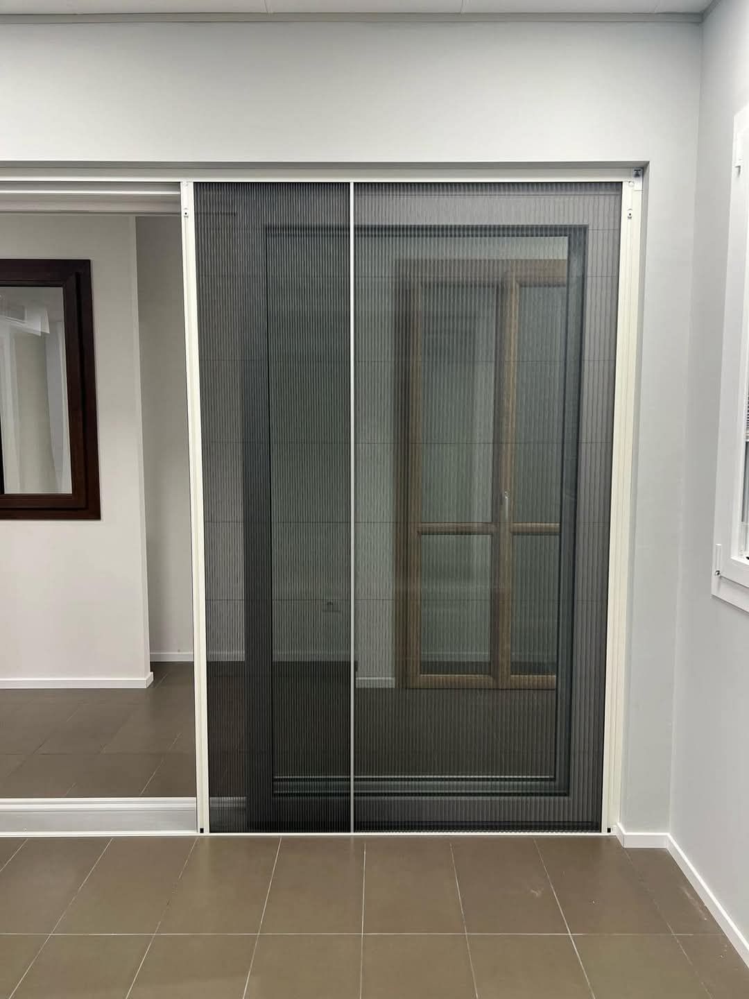 Zanzariera scorrevole plissettata con telaio bianco; porta su una stanza, con finestra sullo sfondo.