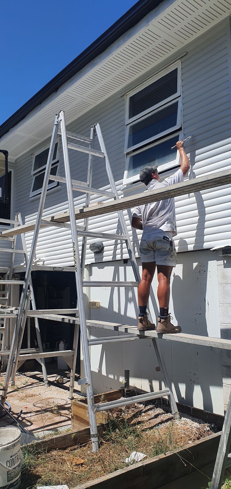 Person Paints Exterior Siding From a Scaffold. Siding is Gray, Sky is Blue — Keppel Coatings in Pacific Heights, QLD