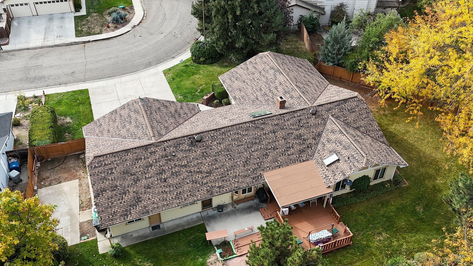 Aerial view of a house with a brown shingle roof, surrounded by green grass and trees with some yellow leaves.