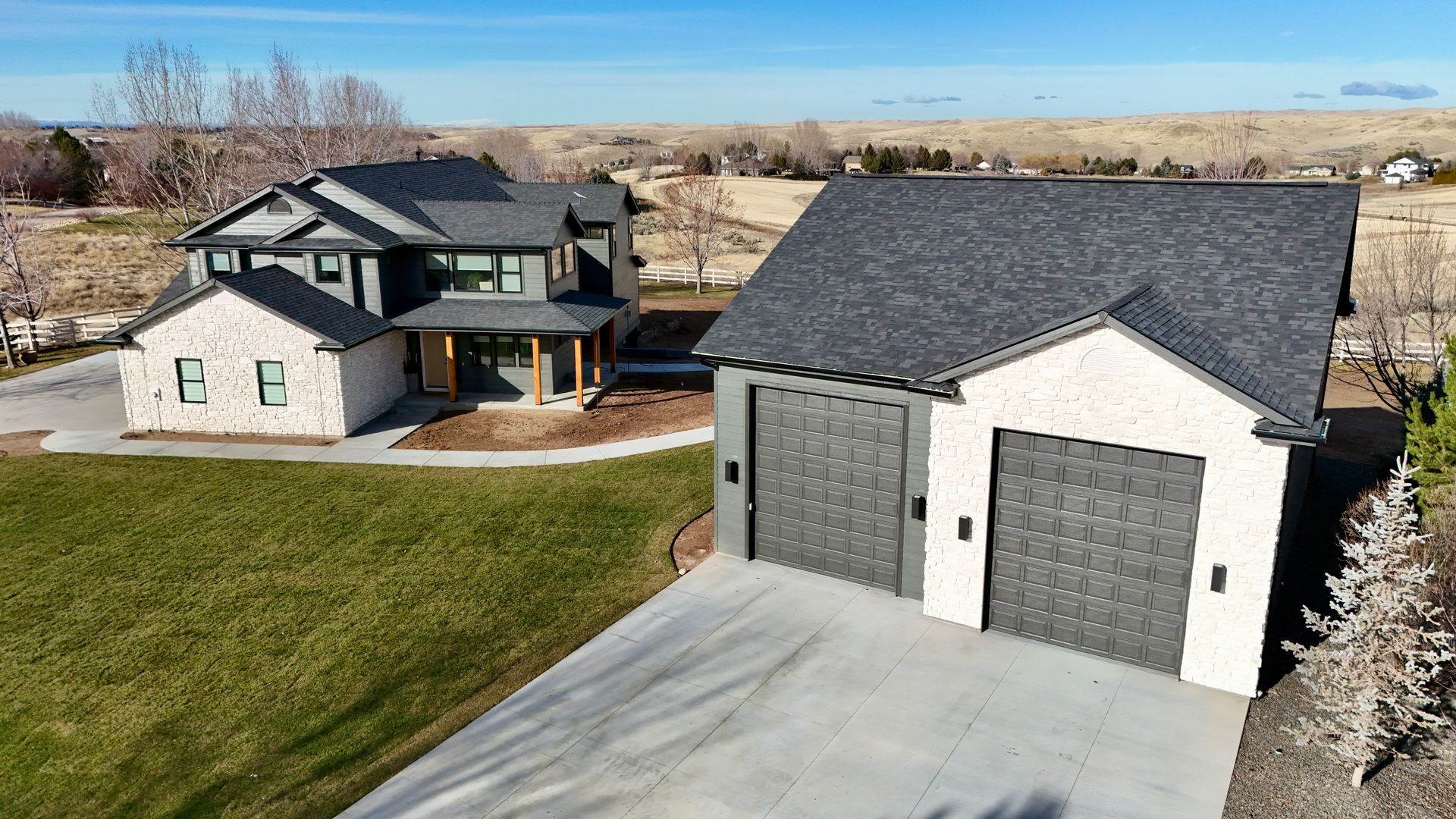 Two-story modern house and detached garage with grey roofs and light brick exterior, on a grassy lot with a driveway, against a blue sky.