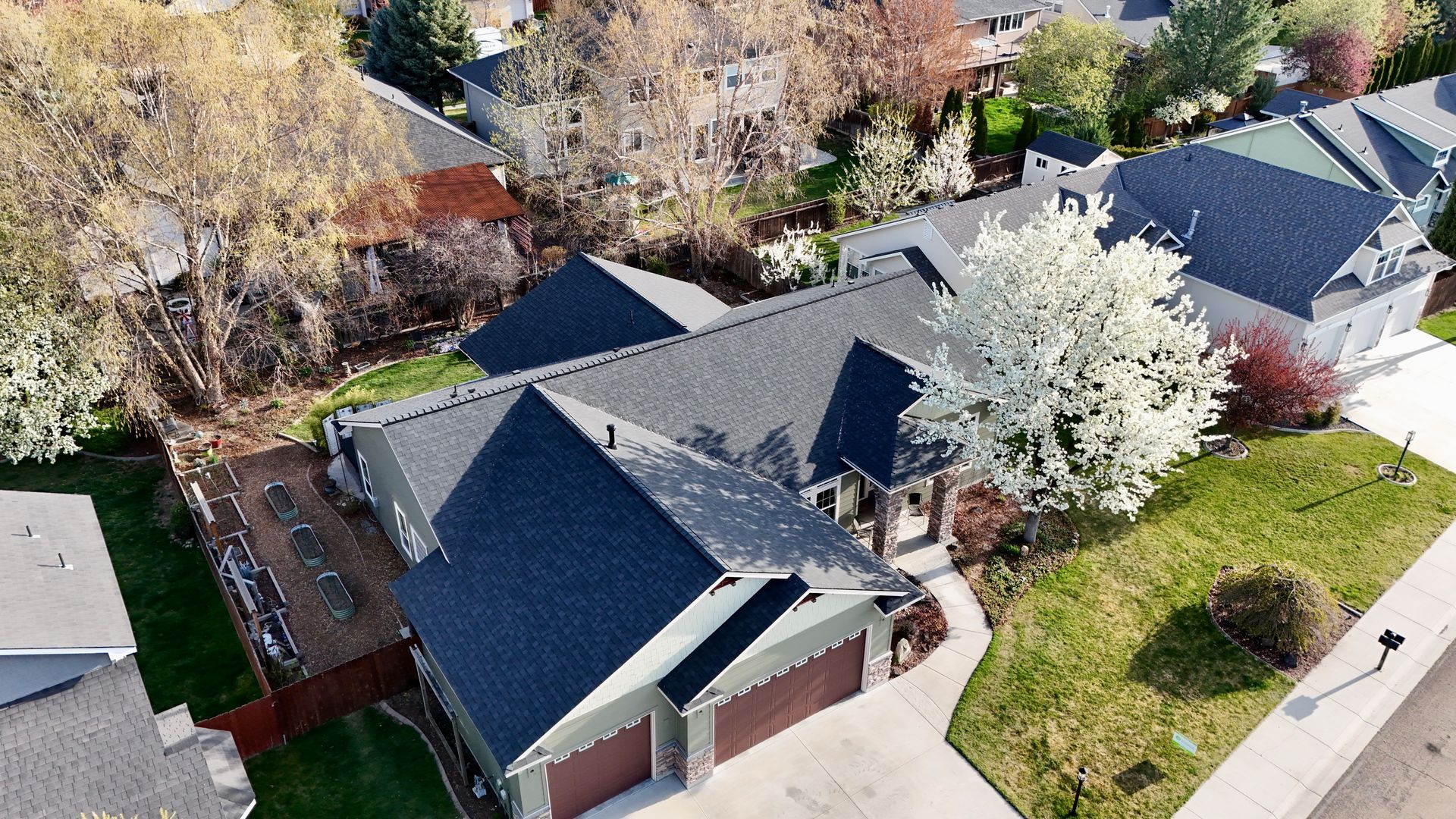 Aerial view of a house with a dark roof, a green lawn, and a white flowering tree in front.  It's located in a residential neighborhood.