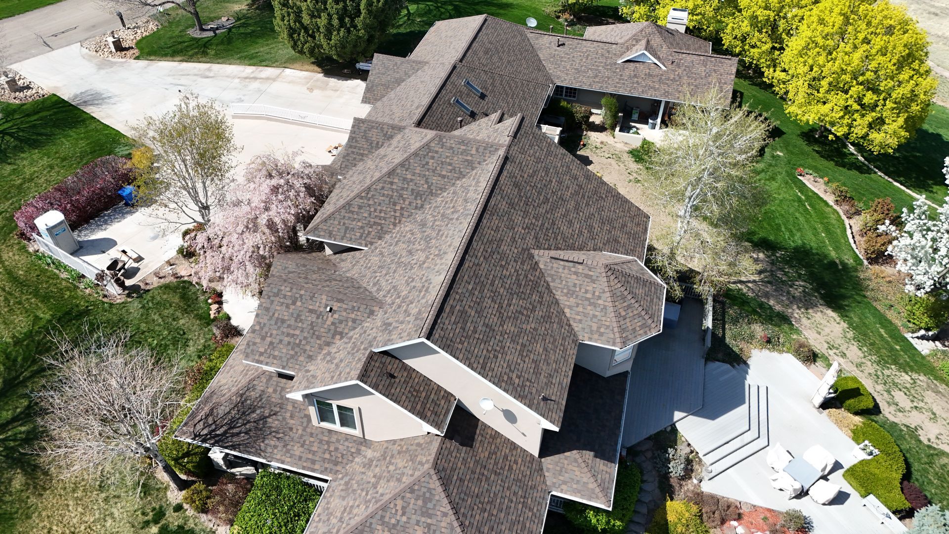 Aerial view of a multi-winged house with a brown shingled roof, surrounded by green lawns, trees, and a long driveway.