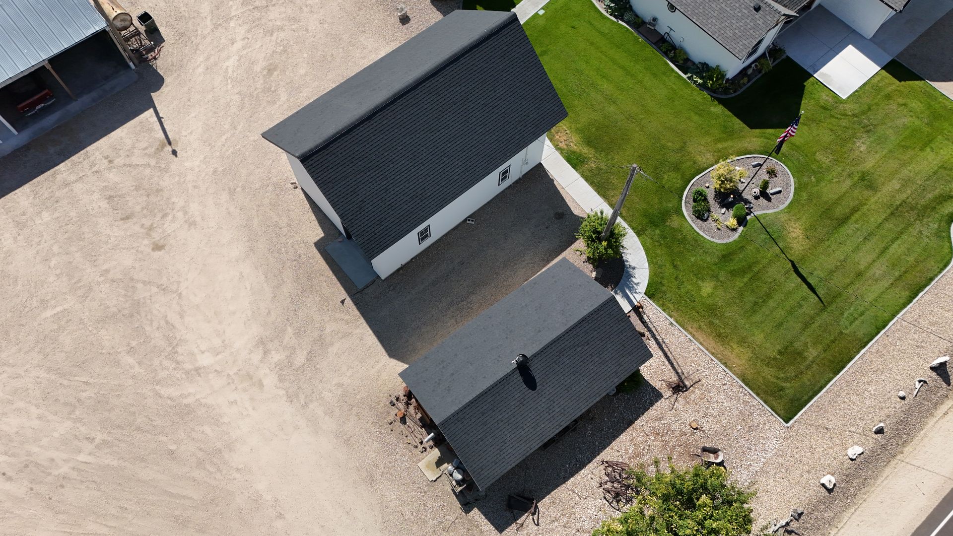 Overhead view of two dark-roofed buildings on a gravel lot, with a green lawn and neighboring houses visible.