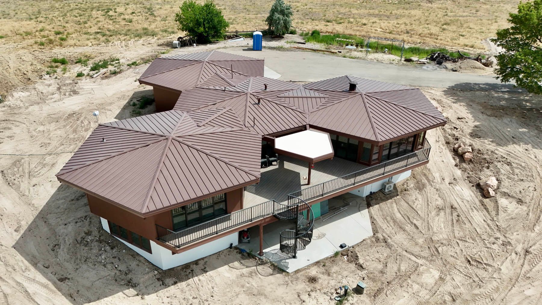 Aerial view of a brown-roofed house with a deck, set on a dirt lot with some vegetation in the background.