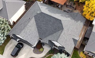 Aerial view of a house with a complex gray roof, driveway with a black car, and surrounding trees.