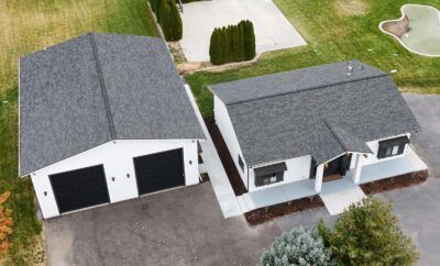 Aerial view of a white house and detached garage with dark grey roofs and black garage doors, set on a green lawn.