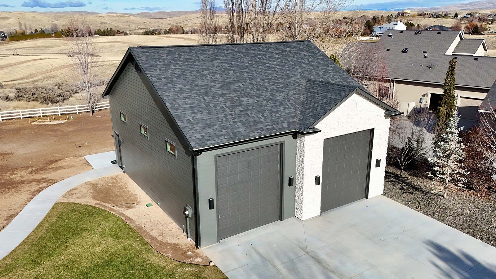 Two-car garage with dark gray siding, light-colored garage doors, and a black asphalt roof. Concrete driveway and green lawn in front.