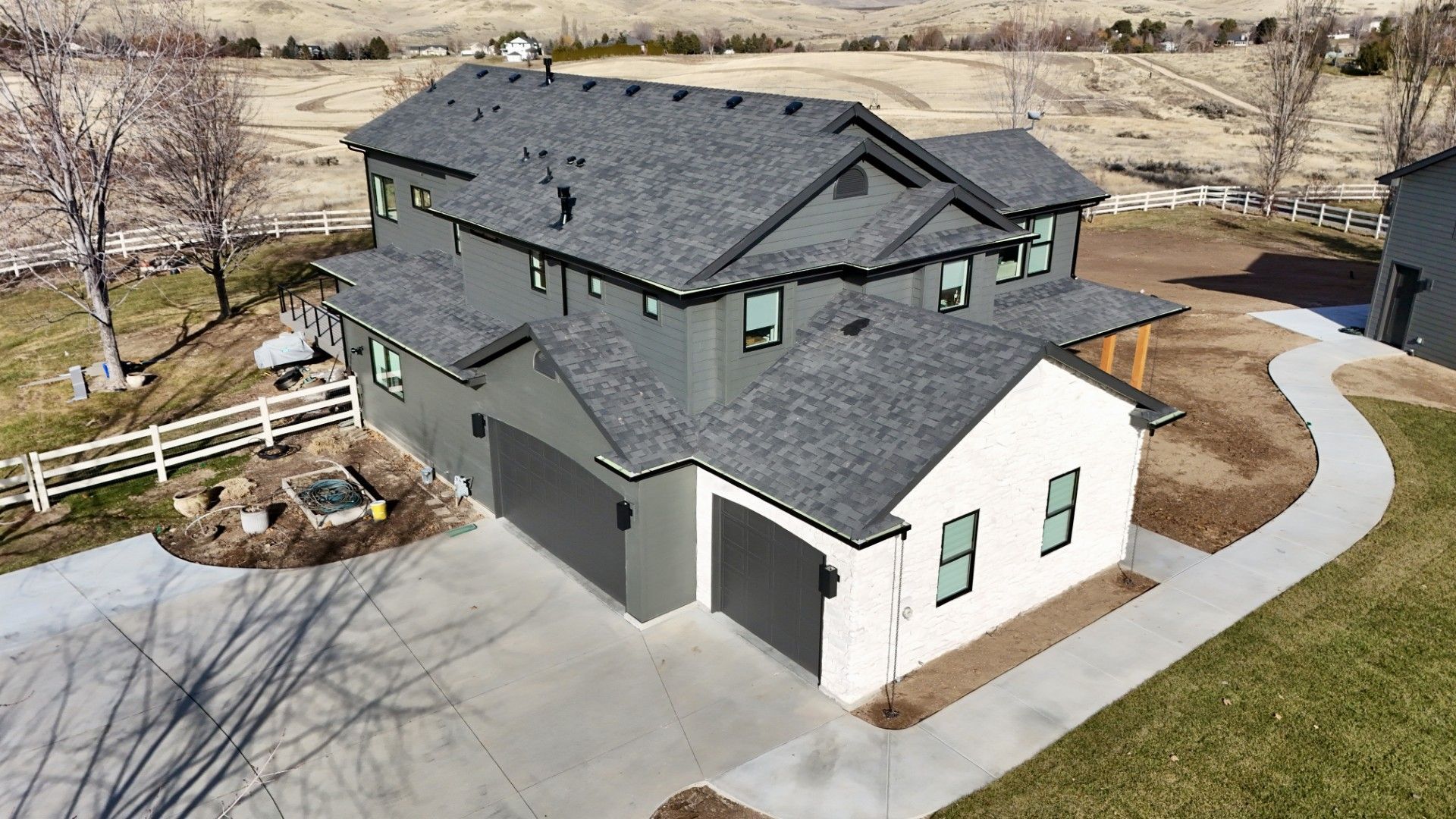 Aerial view of a modern, two-story house with a gray roof and exterior, two garage doors, and a winding driveway.